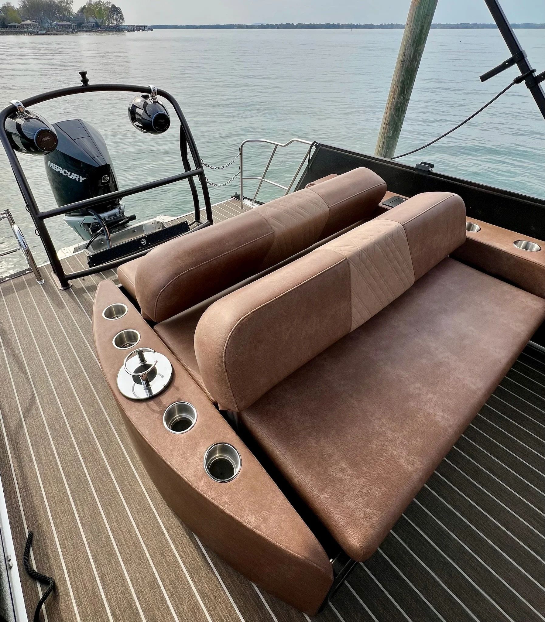 View of a boat's seating area with tan upholstered cushions and built-in cup holders, overlooking a calm body of water with distant trees and houses on the shoreline.