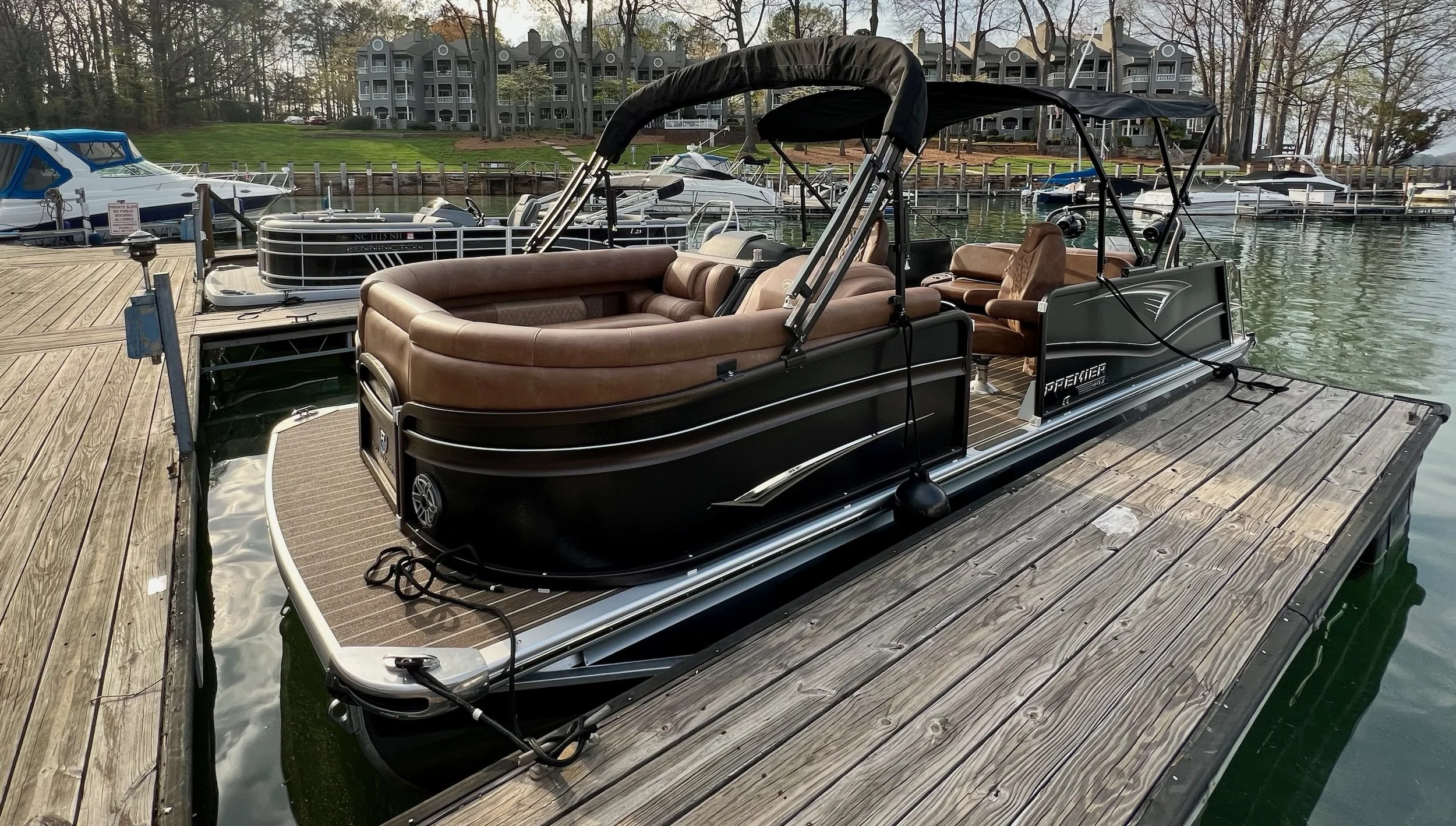 A pontoon boat docked at a marina, featuring brown leather seating and a black exterior, with other boats and water in the background.