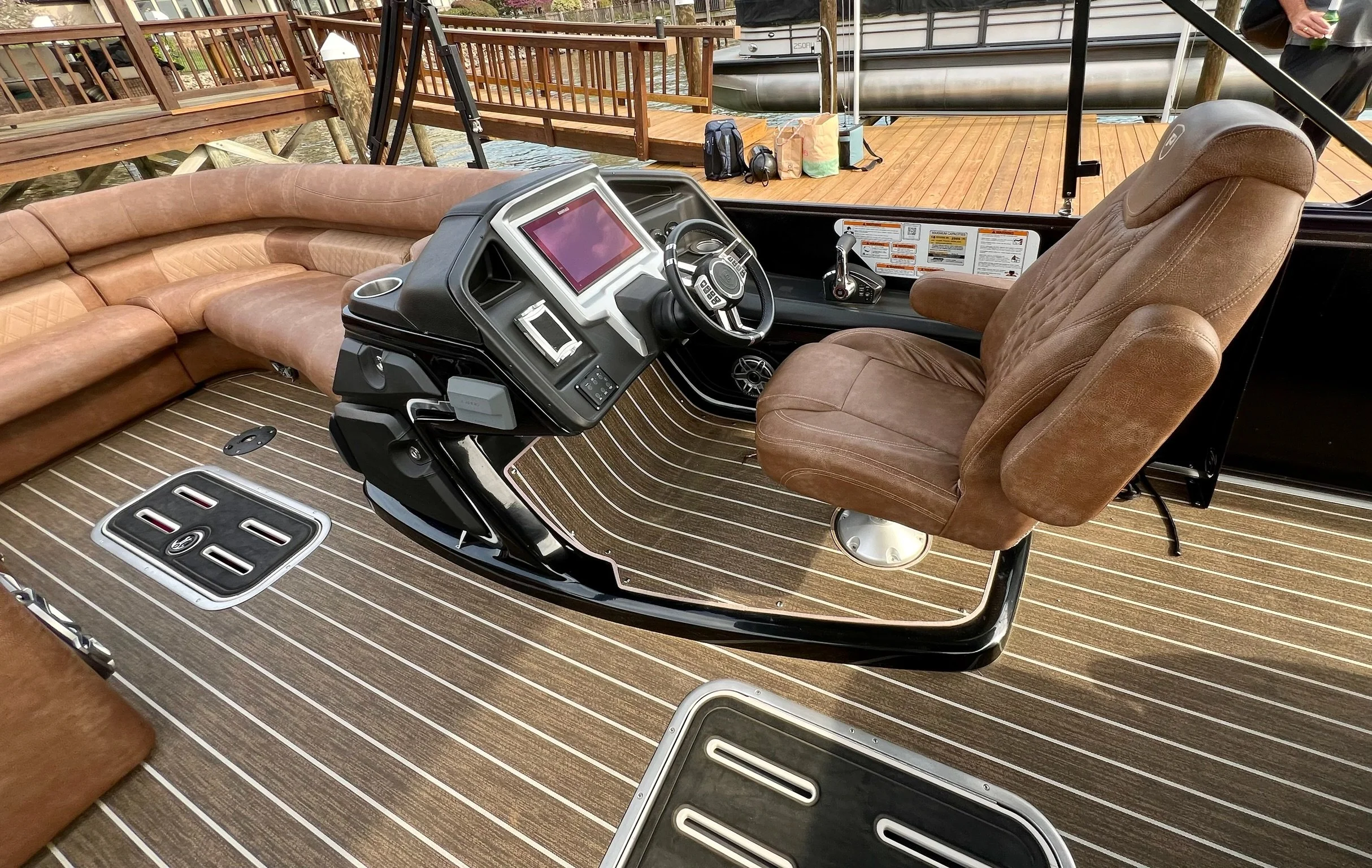 The interior of a boat featuring a captain's chair with brown upholstery, a control console with a steering wheel and display screens, a brown leather couch, a wooden deck floor with striped pattern, and a dock with wooden railings and water in the b