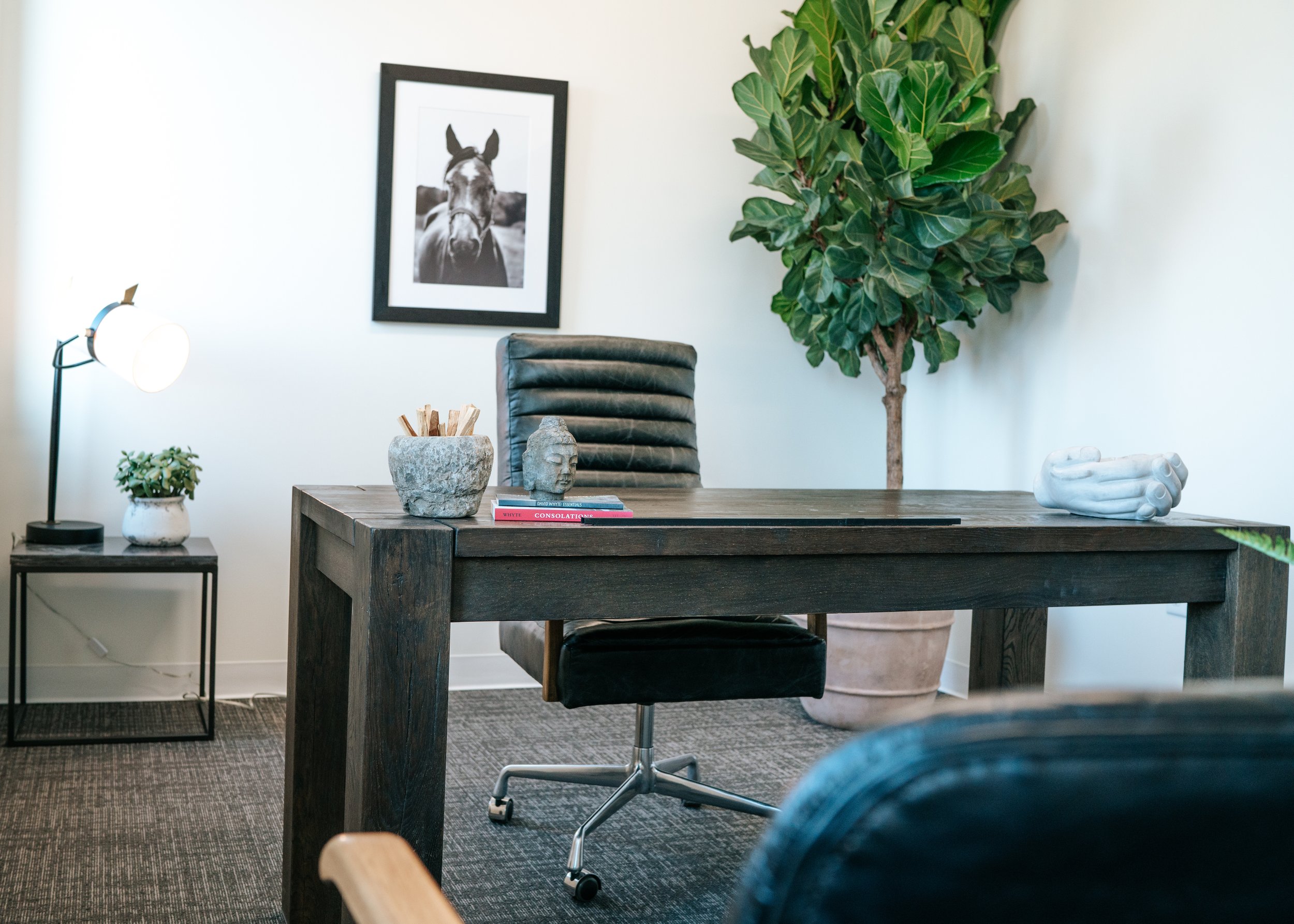 Modern office with a dark wood desk, black leather office chair, potted plant, framed black-and-white horse photo, decorative hand sculpture, books, and a small side table with a lamp and plant.