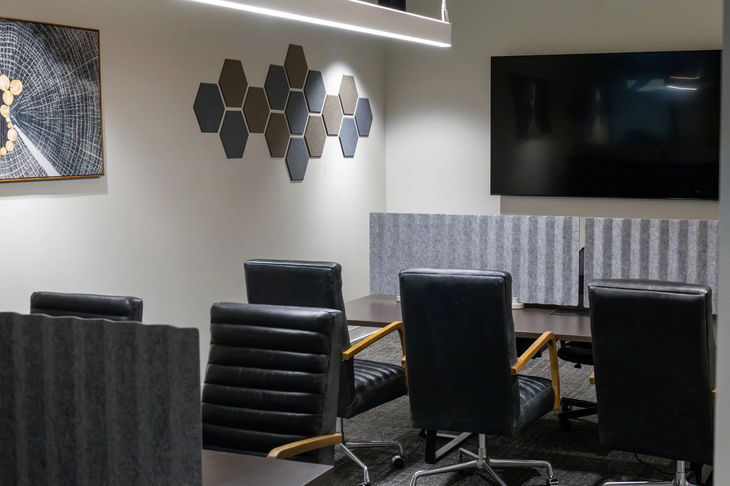 Empty conference room with black leather chairs, a large mounted TV screen, and wall decor including a framed abstract artwork and hexagonal soundproof panels.