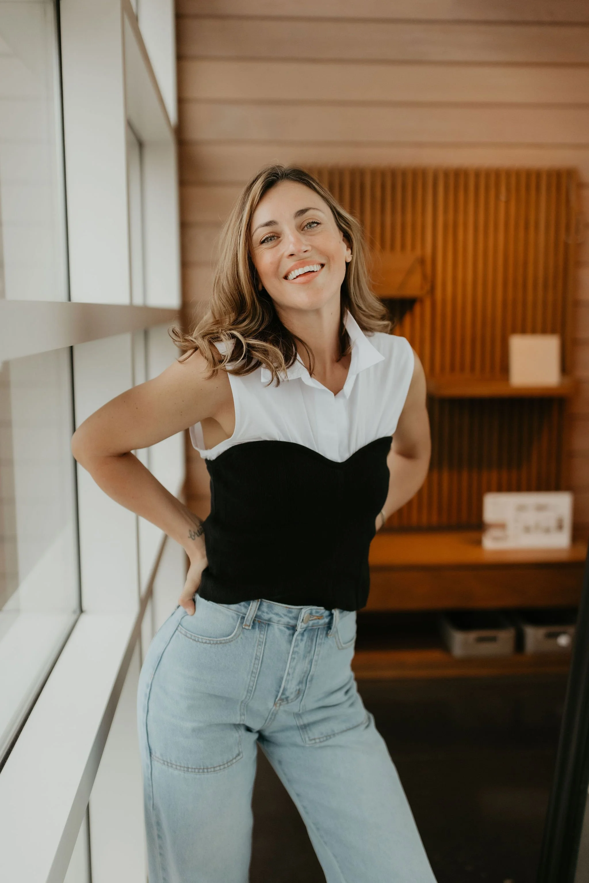 A woman with shoulder-length, wavy brown hair smiling and leaning against a white window sill with hands on her hips, standing indoors.