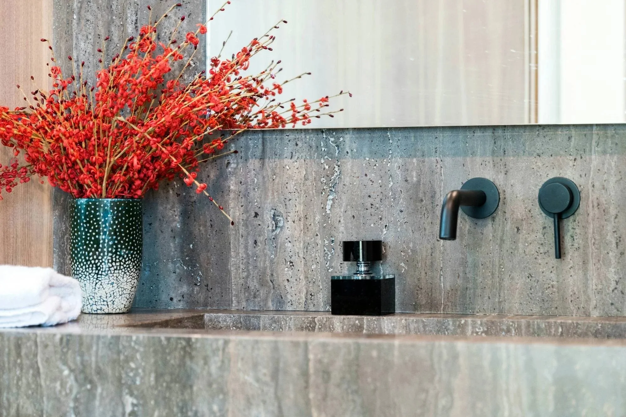 Indoor bathroom sink area with a large decorative vase containing red dried flowers, a black soap dispenser, and a black faucet on a gray marble countertop with a matching backsplash.
