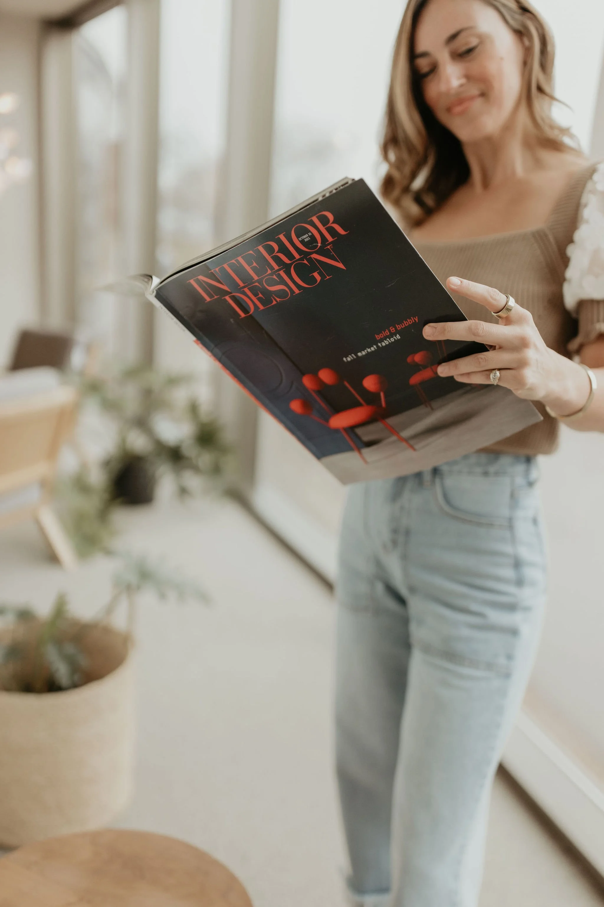 A woman reading a magazine titled 'Interior Design' in a well-lit room with large windows.