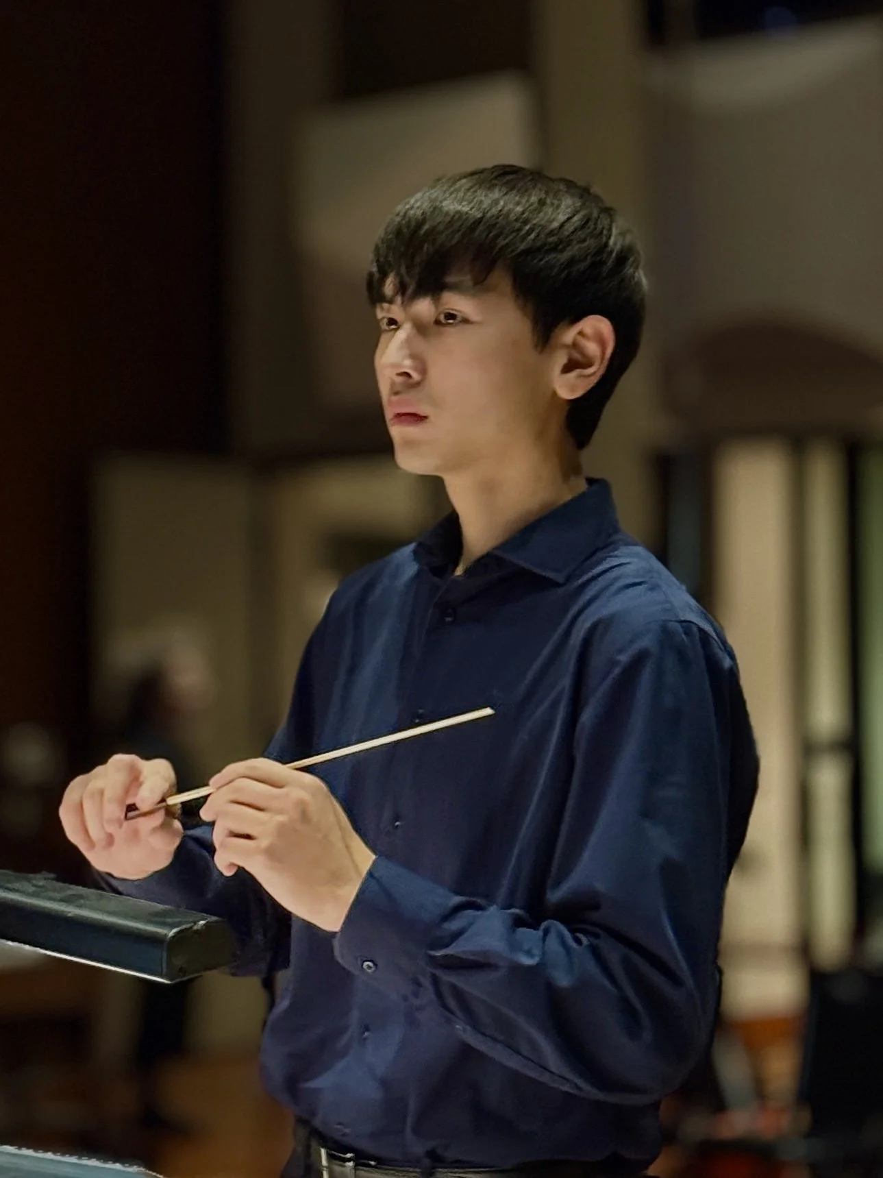 A young man with dark hair wearing a navy blue shirt, holding a conducting baton, conducting at Warner Bros. Studio