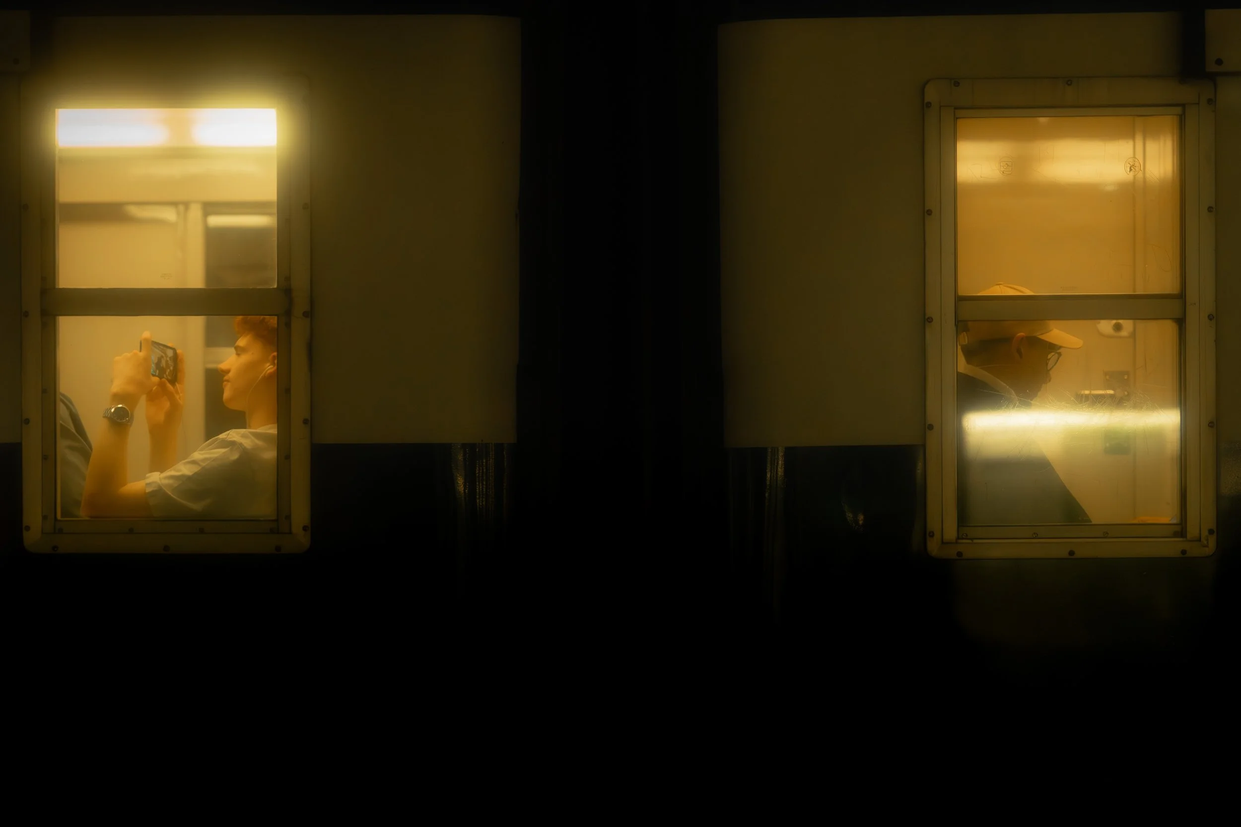 Two people seen through separate windowpanes, one on the left taking a photo or video with a phone, the other on the right wearing a hat and glasses, inside a dimly lit room.