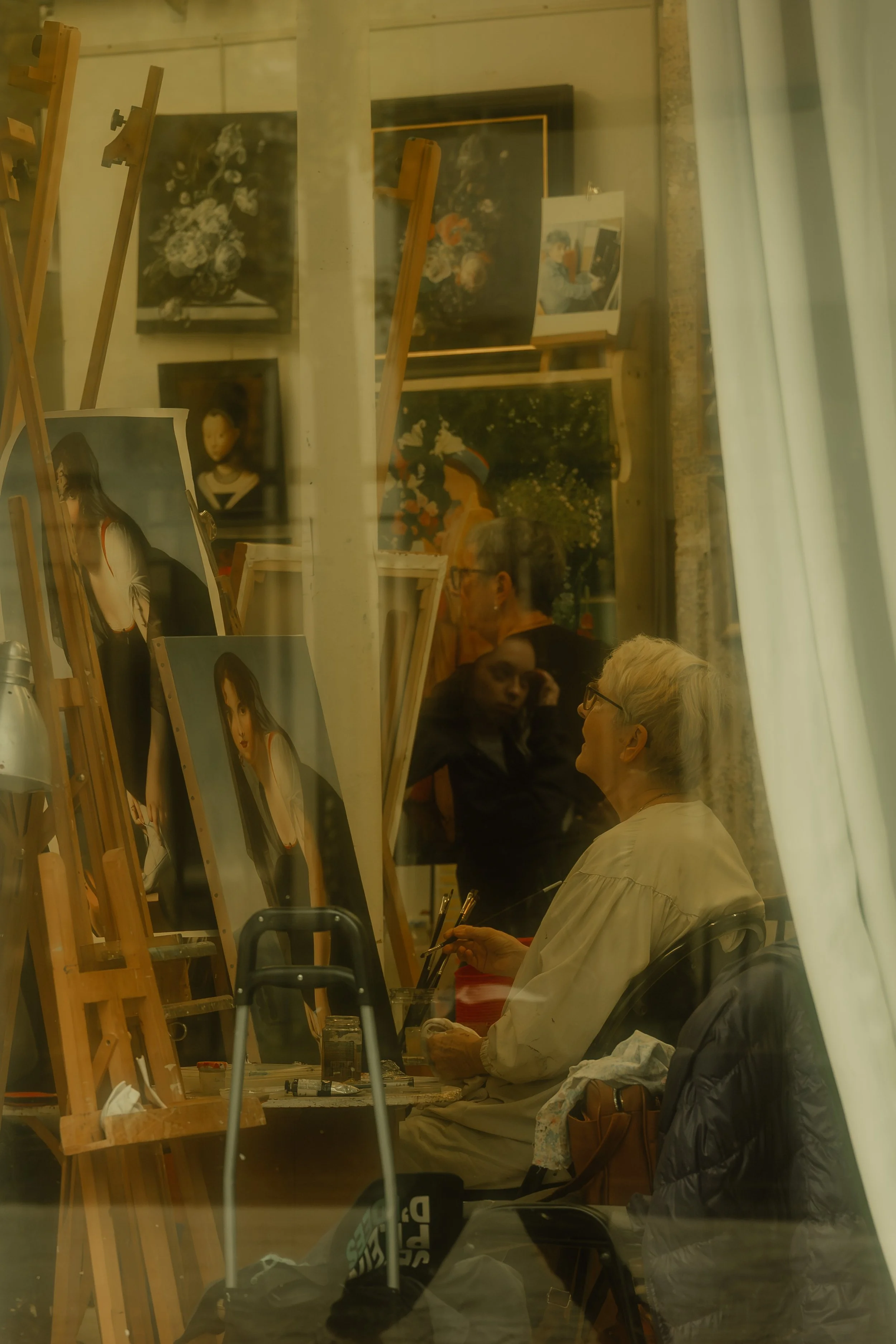 An elderly woman with short white hair and glasses, painting in an art studio, surrounded by portraits and still life paintings on easels and walls, with two other people visible in the background.