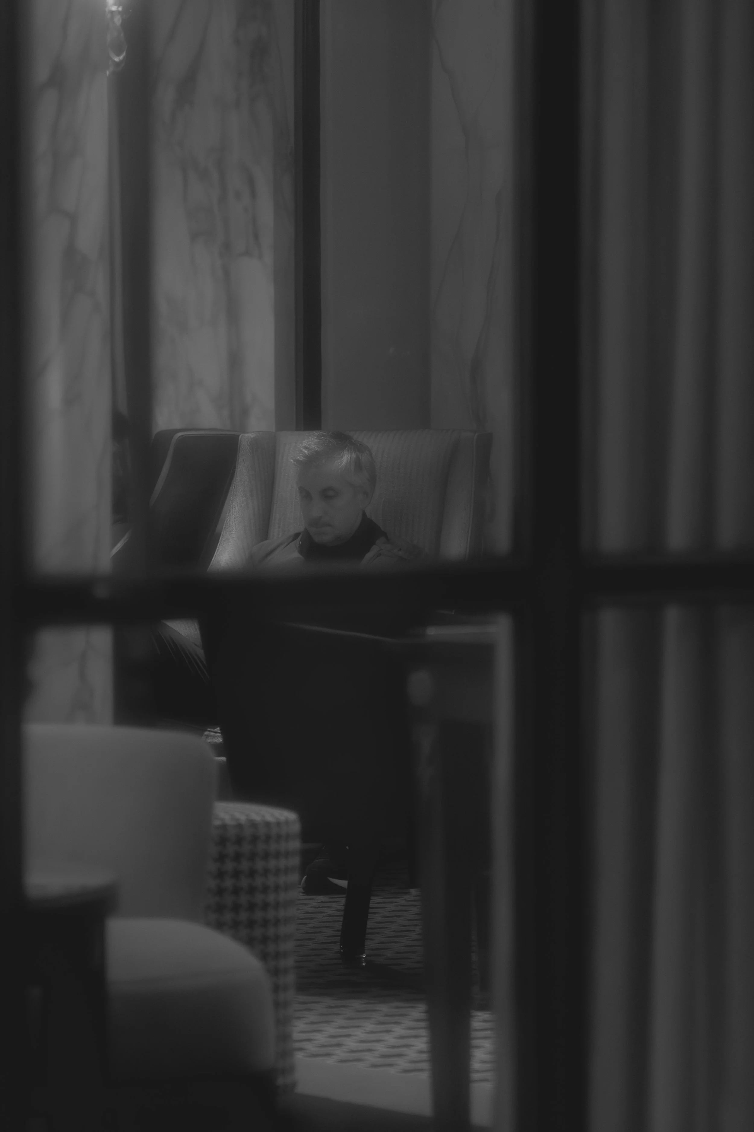 A man sitting alone in a lounge, seen through a window with multiple panes, in black and white.