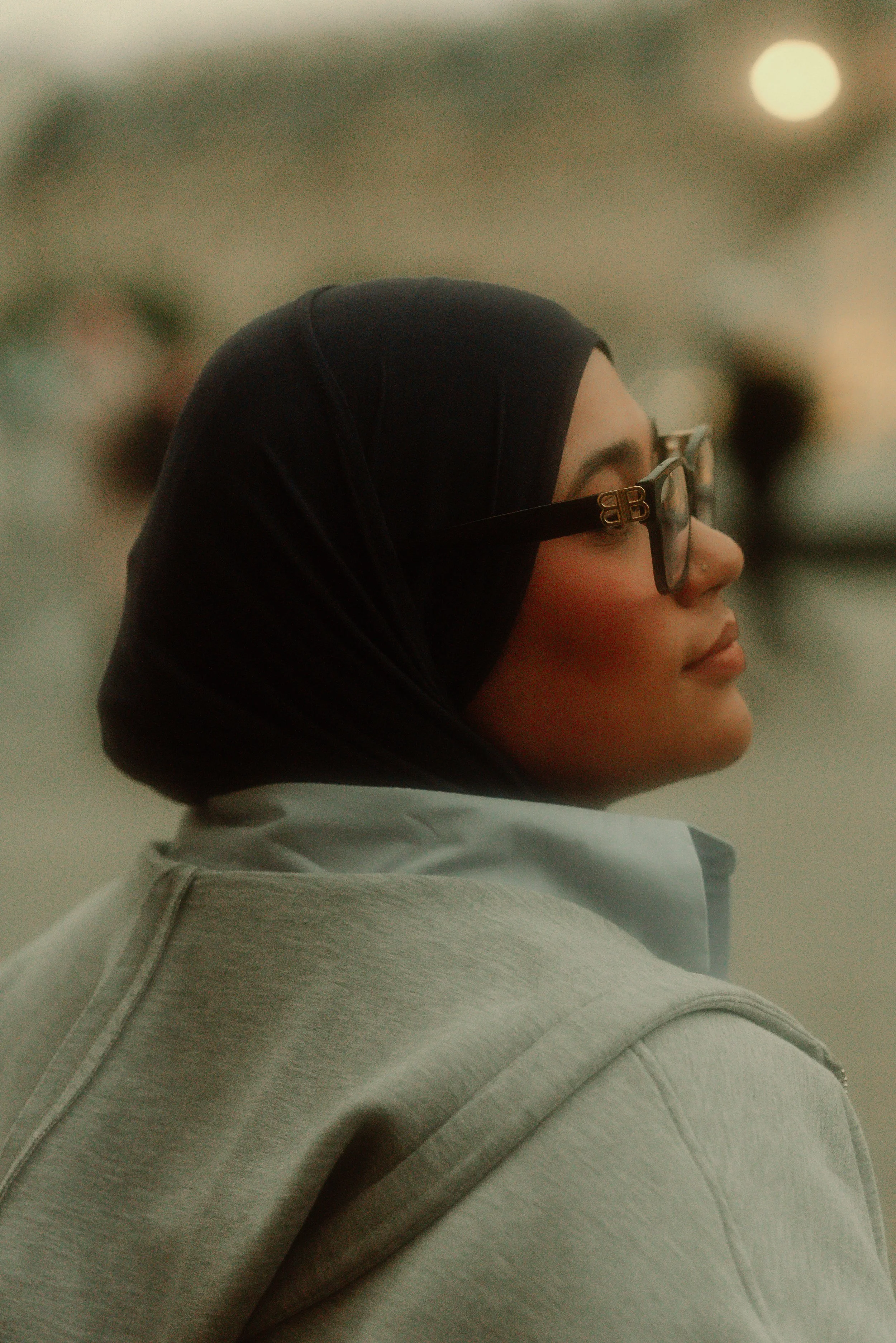 Profile of a woman wearing glasses, a black headscarf, a white hoodie, and a blue shirt outdoors at dusk.