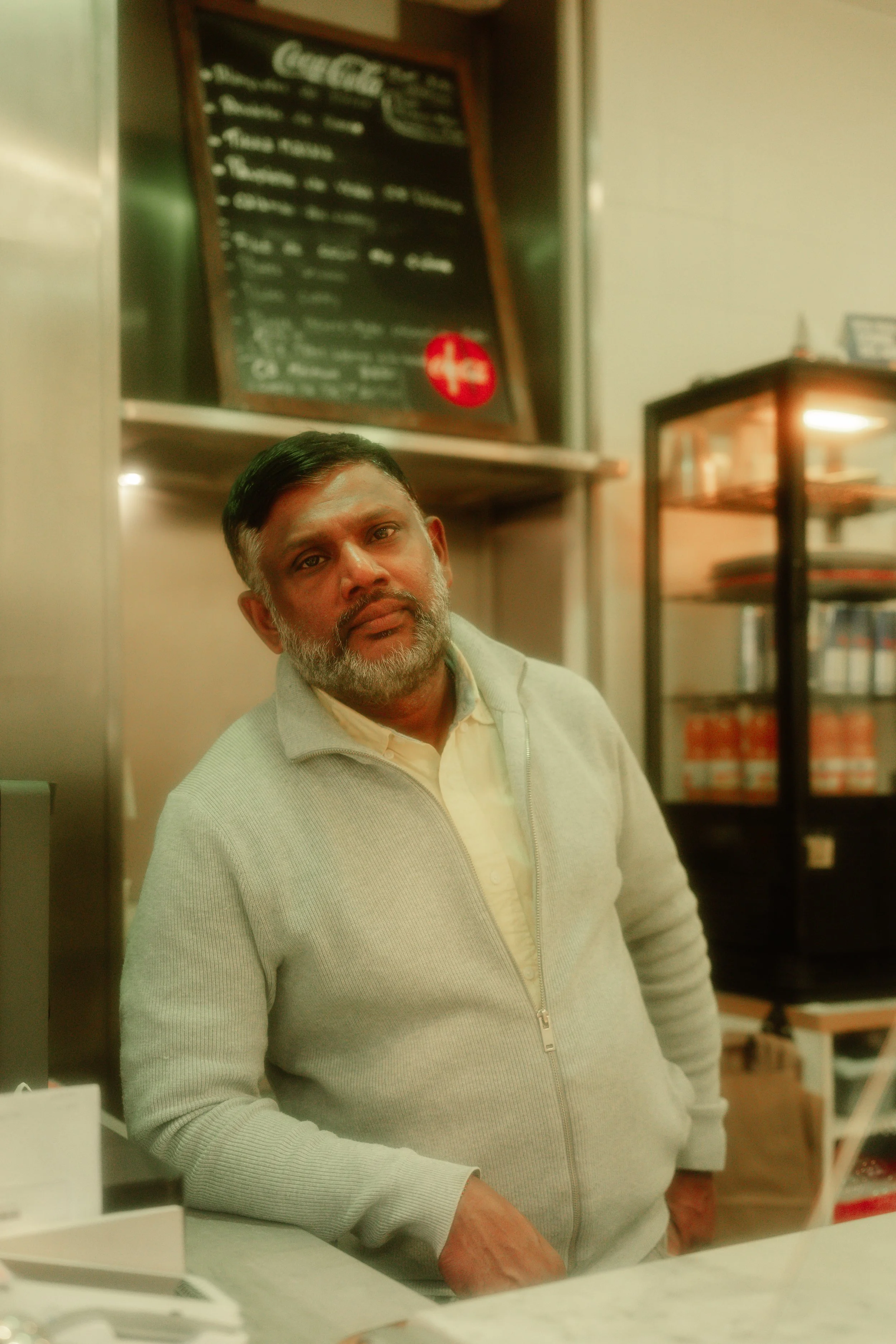 A man with a beard and mustache leaning against a counter in a cafe or restaurant with a menu board and shelves in the background.