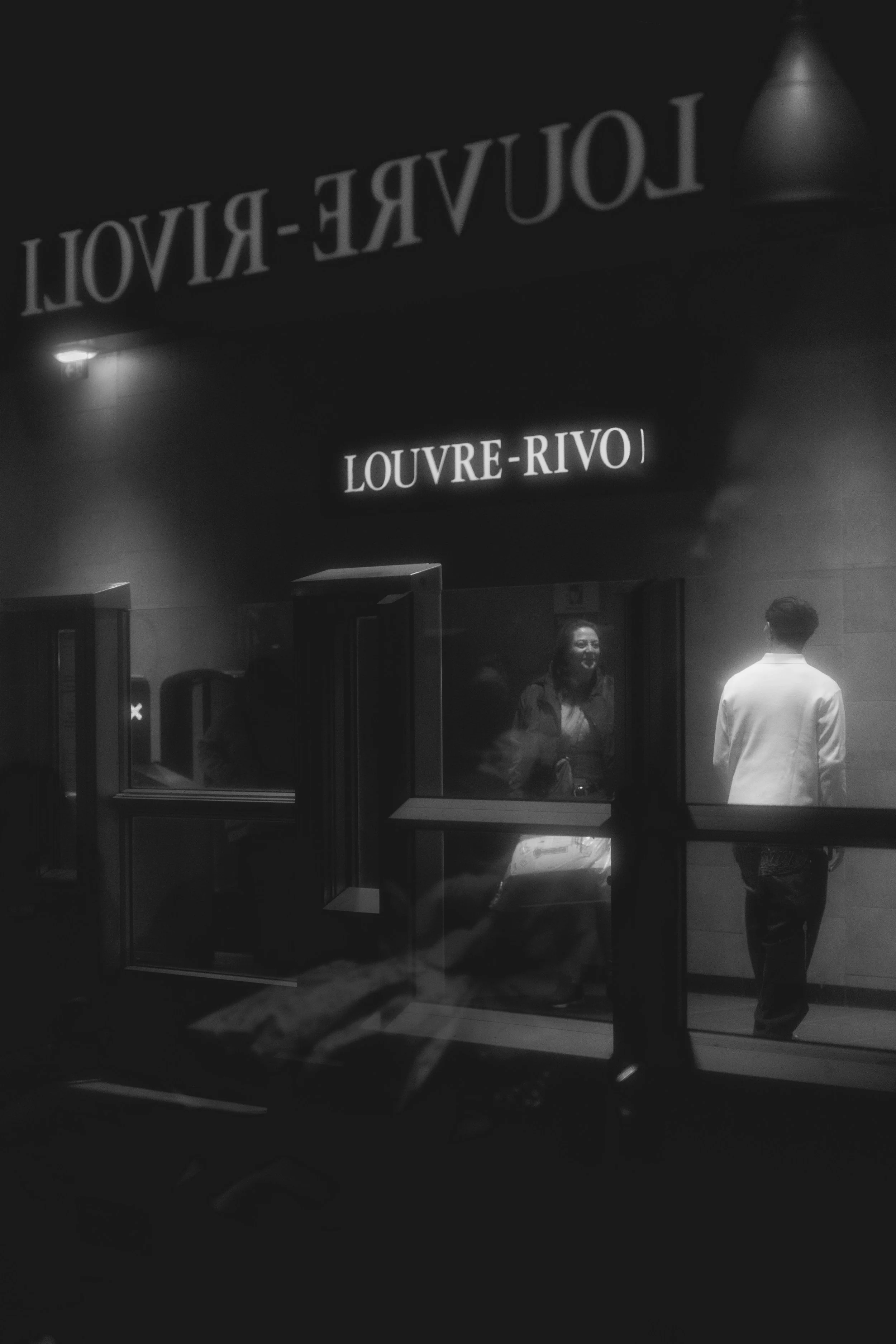 Night view of Louvre-Rivoli entrance, with two people inside near the window, one smiling woman and one man with back turned, with illuminated signs and glass reflections.