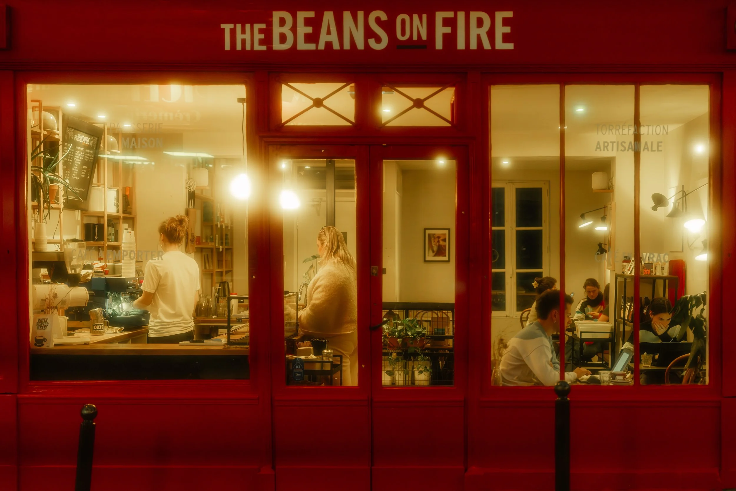 Inside a cozy coffee shop named 'The Beans on Fire' with customers working on laptops and baristas preparing drinks, illuminated by warm indoor lighting.