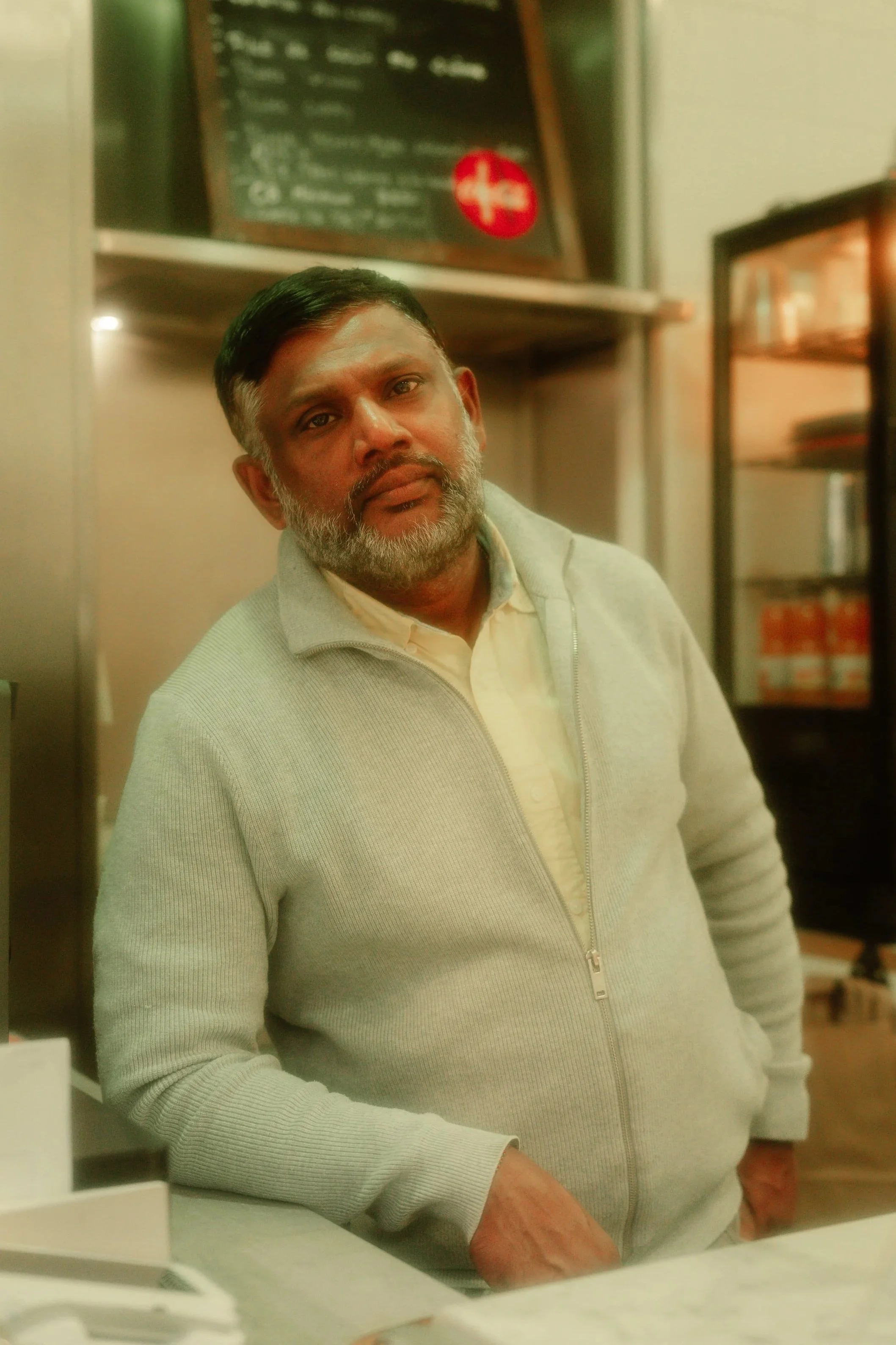 A middle-aged man with a beard, wearing a light gray zip-up sweater and a collared shirt, standing in a cafe or restaurant. There is a blackboard menu in the background and a refrigerated display case.