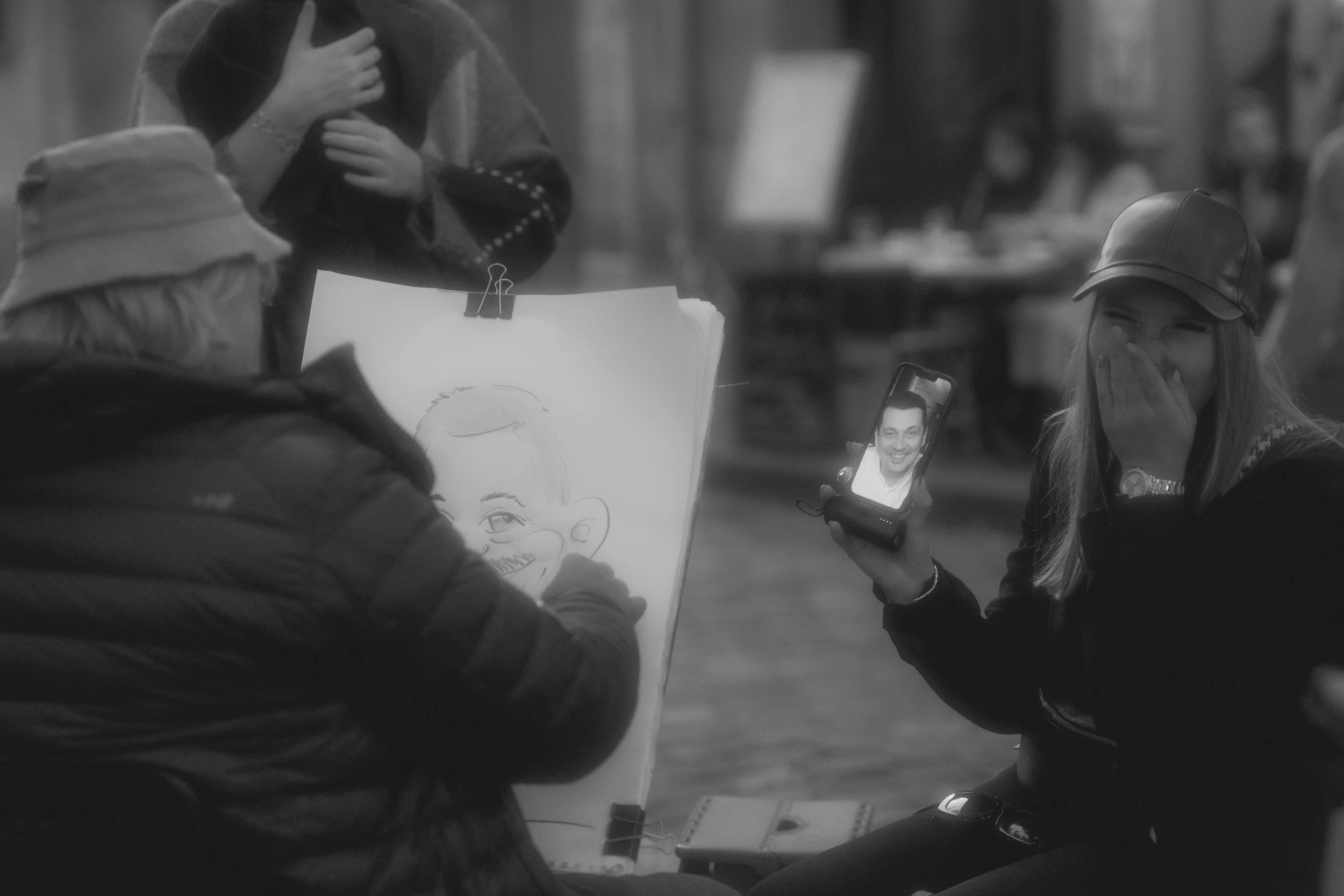 A person is drawing a caricature of a woman who is sitting with her hand over her mouth and smiling, while she is on a video call with someone on her phone, also smiling.