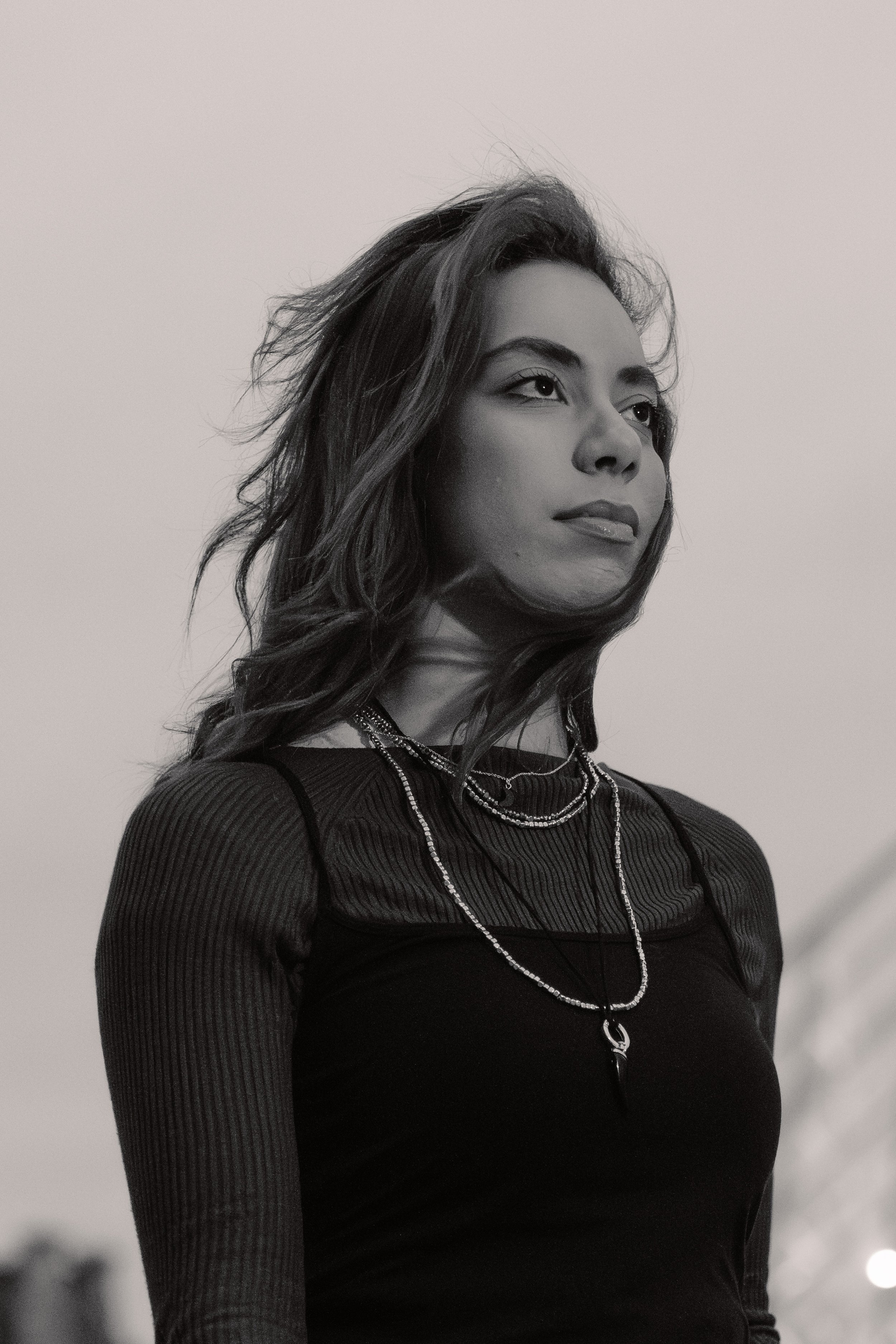Black and white portrait of a young woman with wavy hair wearing layered necklaces and a dark, fitted top.