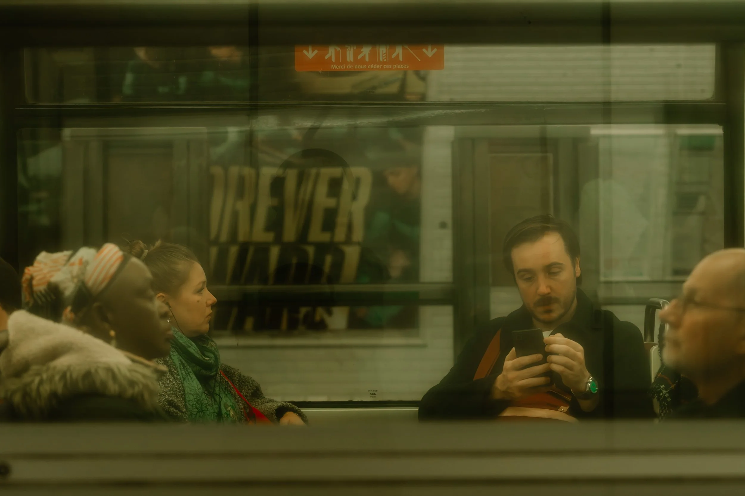People sitting on a subway train, visible through a window, with a man looking at his phone and others with closed eyes or listening to music.