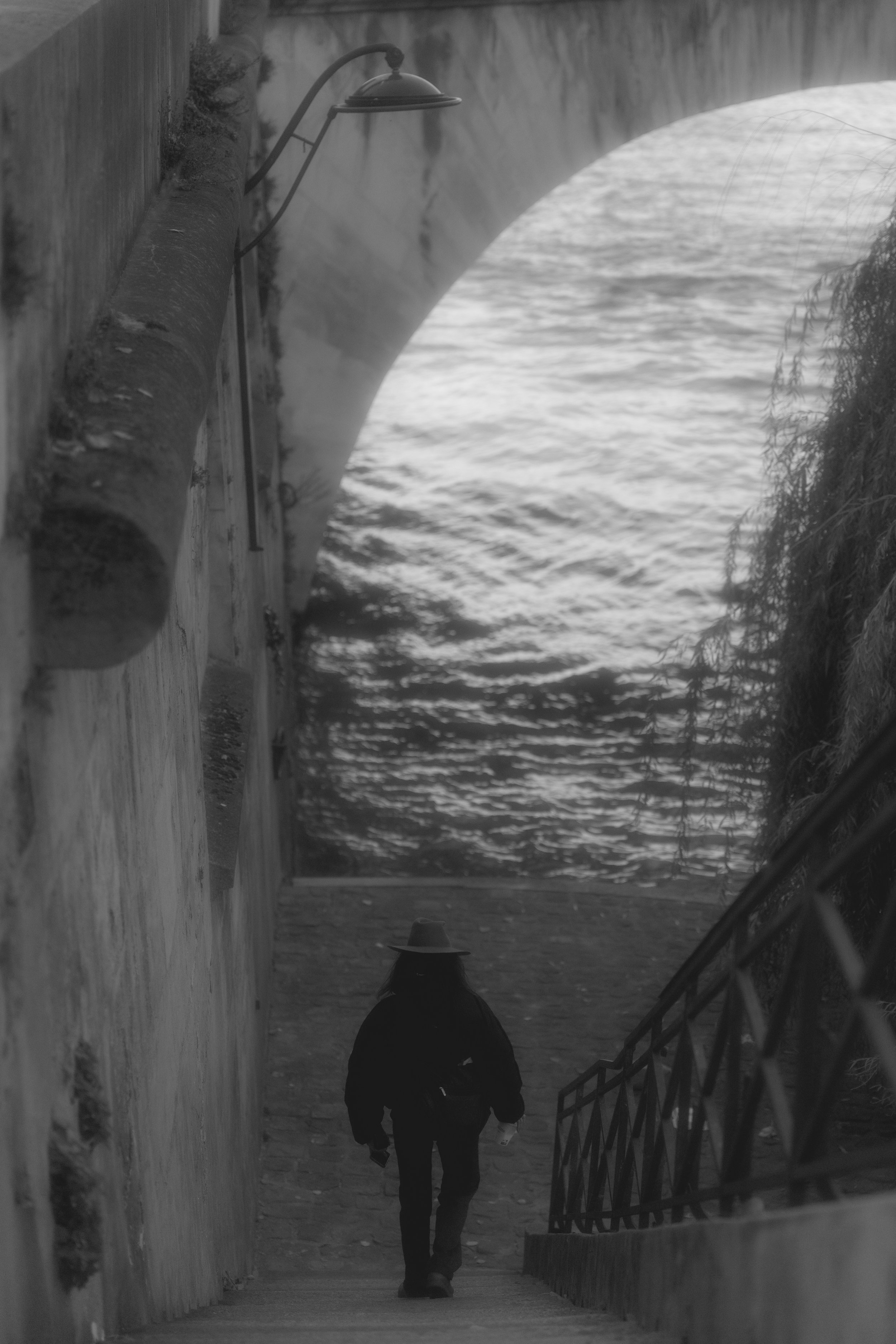 Silhouette of a person wearing a hat walking down stairs beside a waterway with a stone arch bridge overhead.