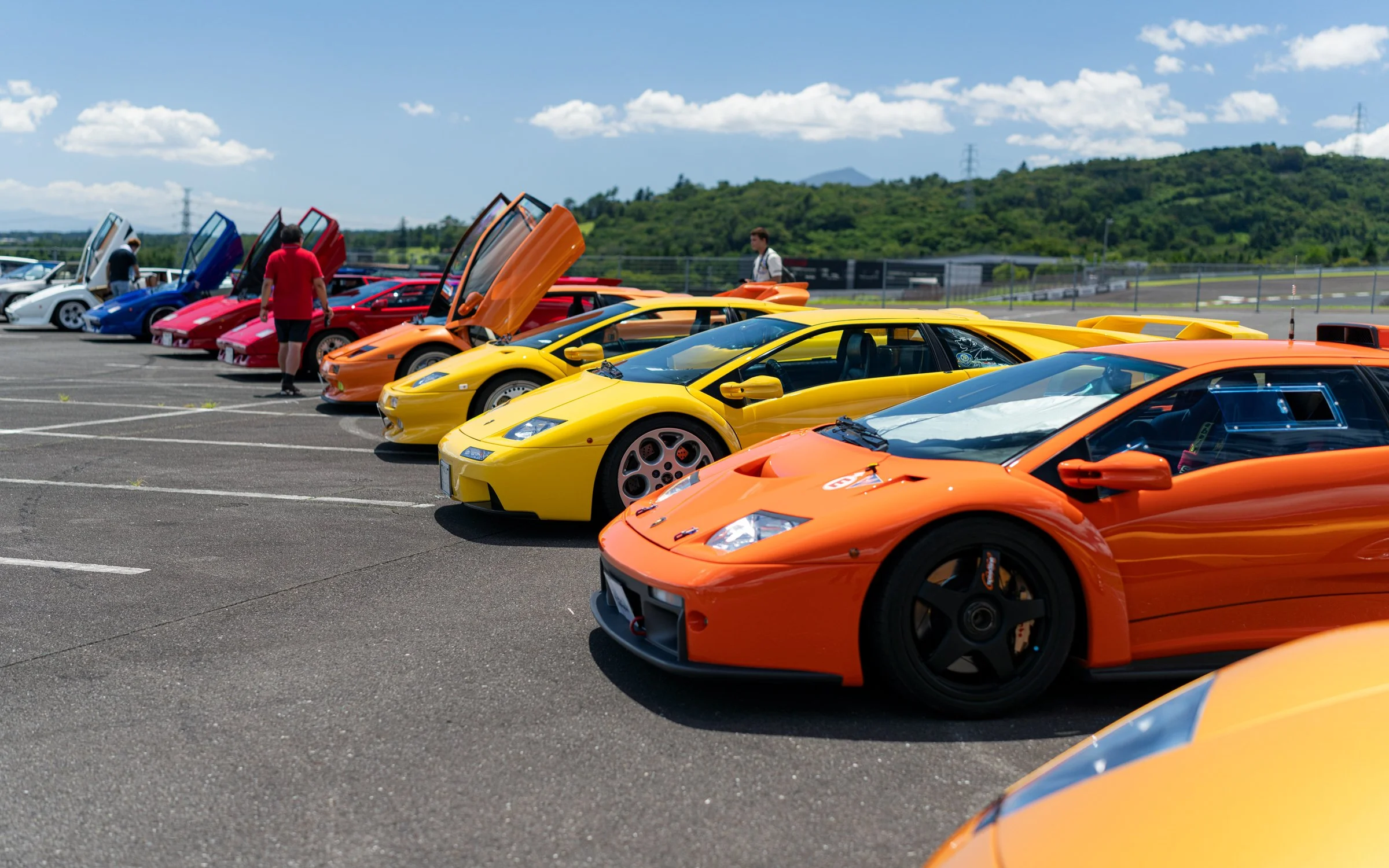 Lineup of colorful sports cars, mostly yellow and orange, with some red, blue, and white, parked with open doors and hoods, at a racetrack on a sunny day with a few people walking around.