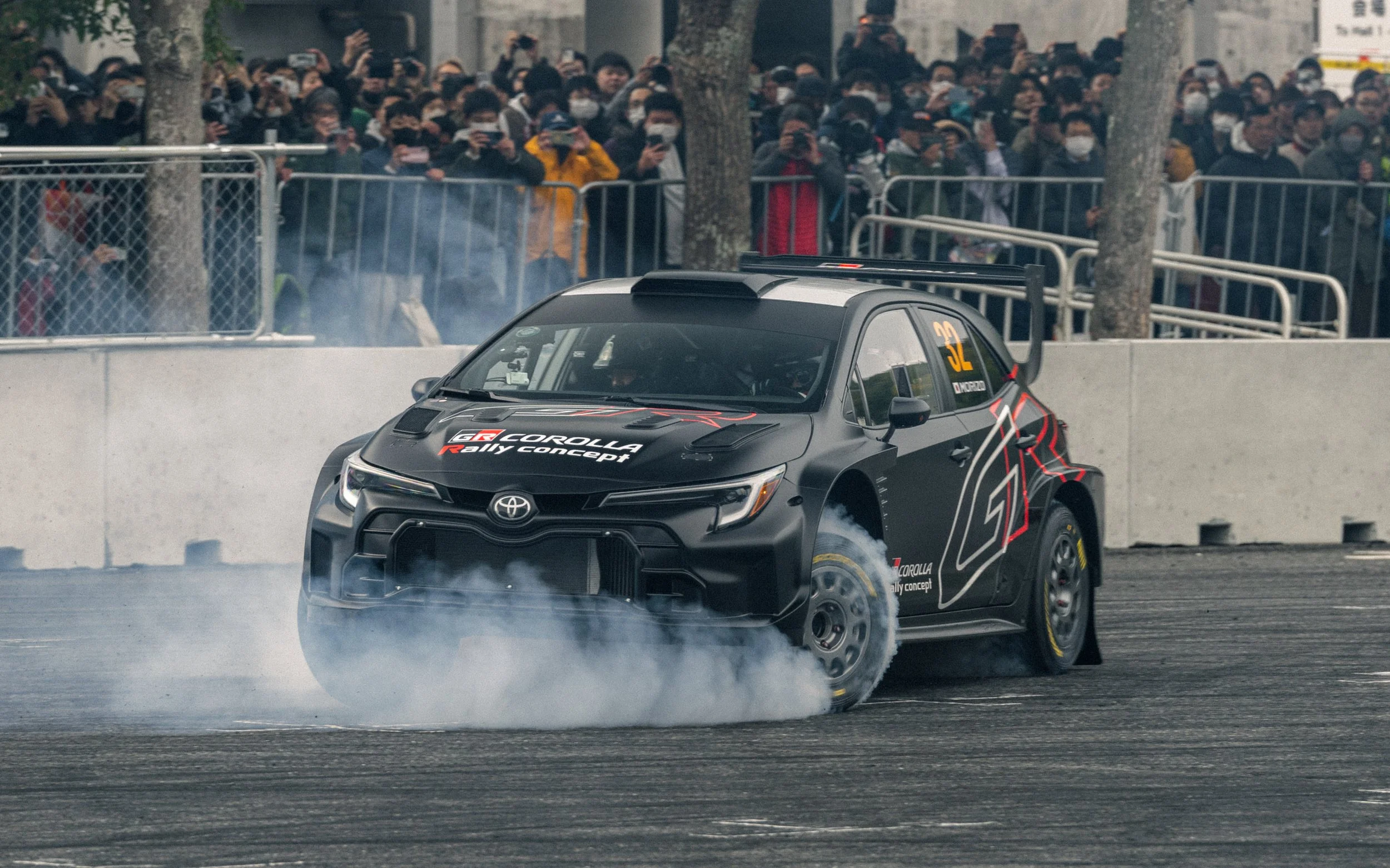 A black Toyota GR Corolla rally car performing a burnout on a racetrack, with smoke coming from the rear tires, and a large crowd of spectators watching behind a fence.