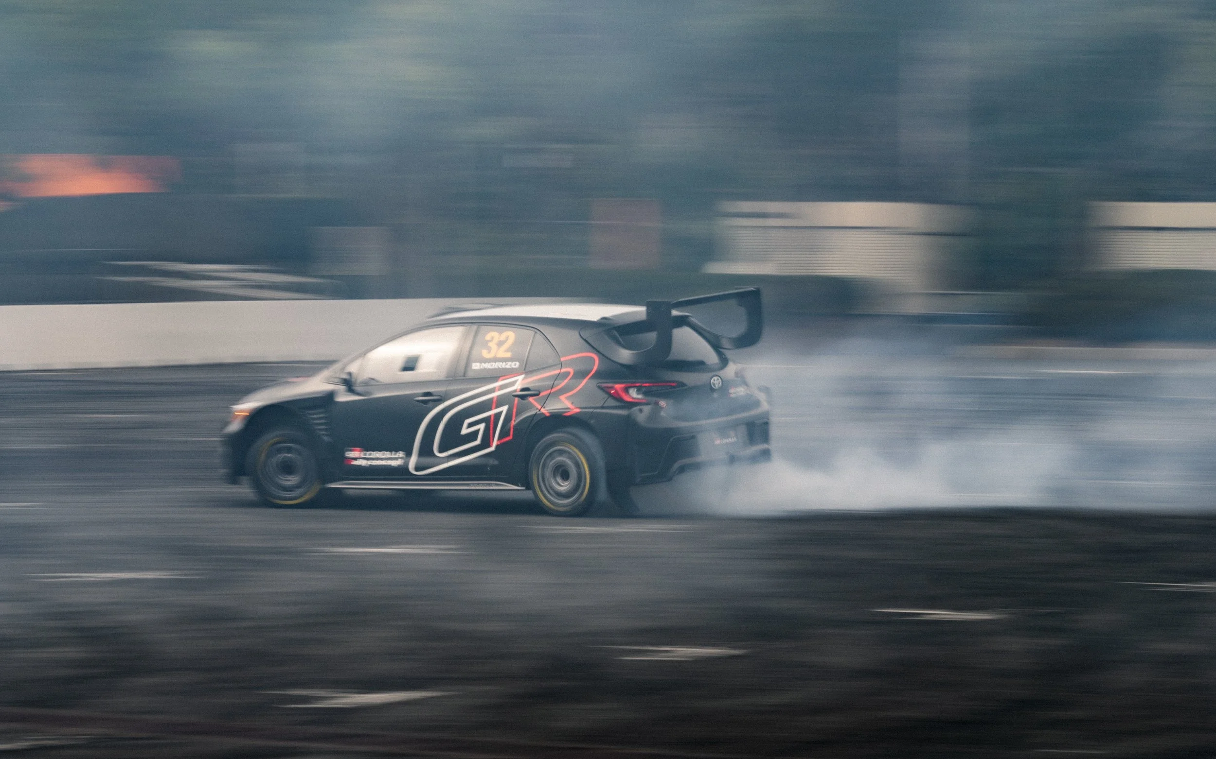 A black race car with a large rear wing, numbered 32, drifting on a race track with smoke trailing from the tires.