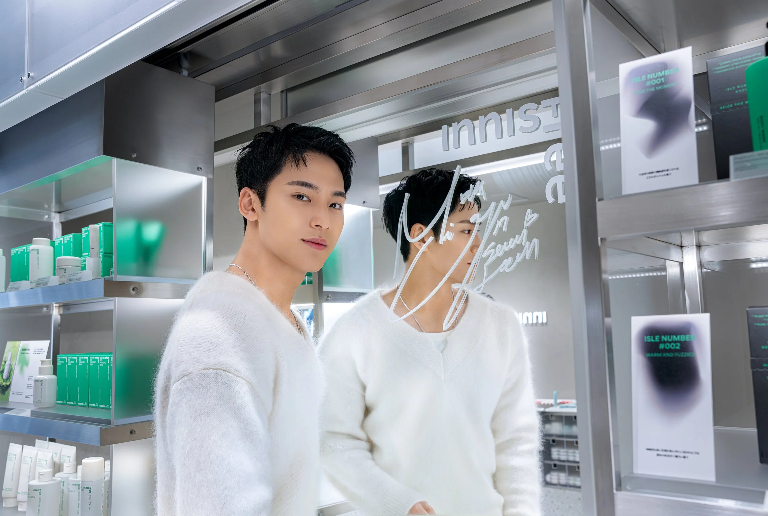 Two young men with black hair and fair skin wearing white sweaters in a store aisle with shelves of skincare products. One man faces the camera, the other is looking at the products. There are fluorescent lights and store signage on the glass mirror.