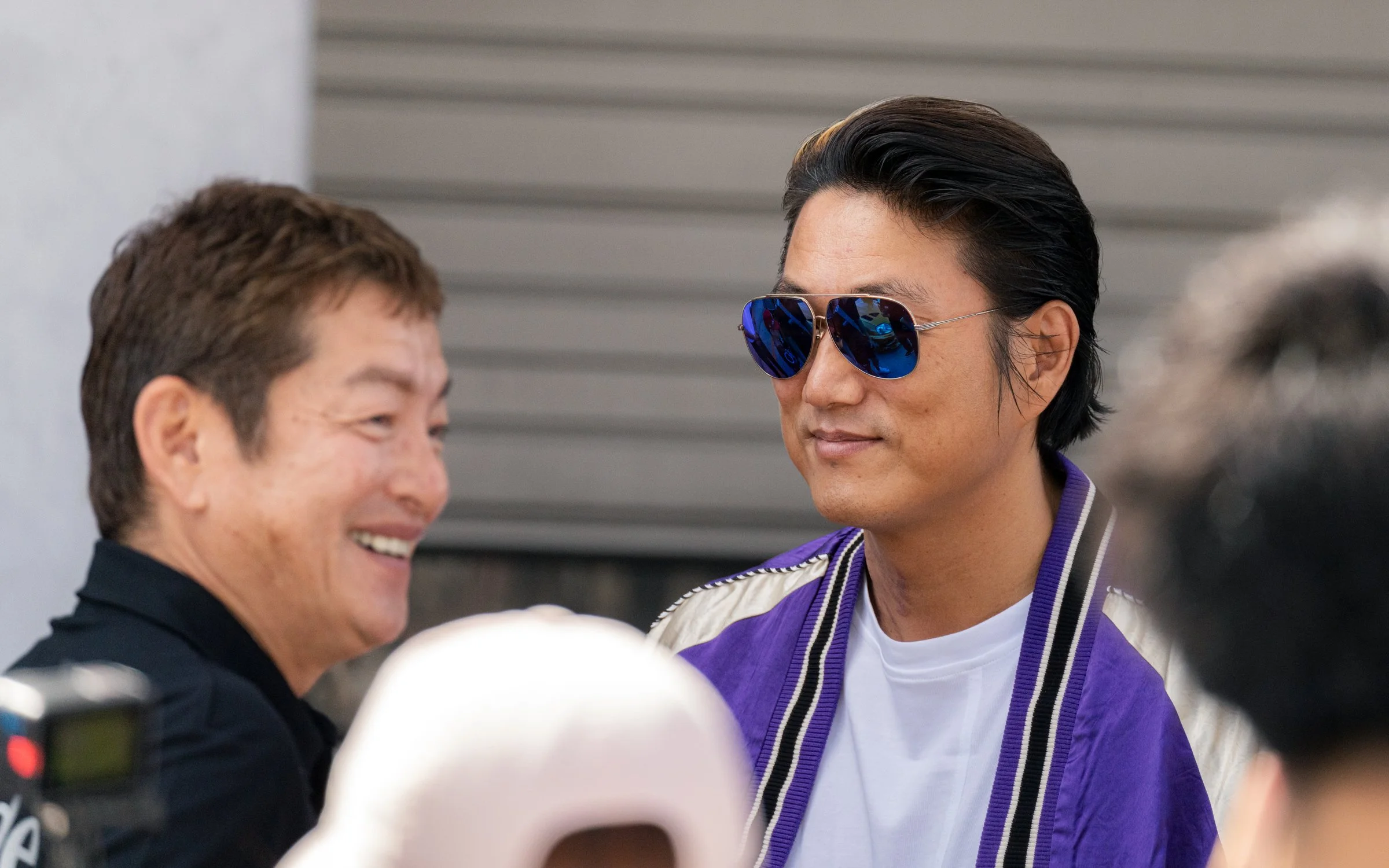 Two men talking, one smiling with show and brown hair, the other wearing sunglasses and a purple jacket.