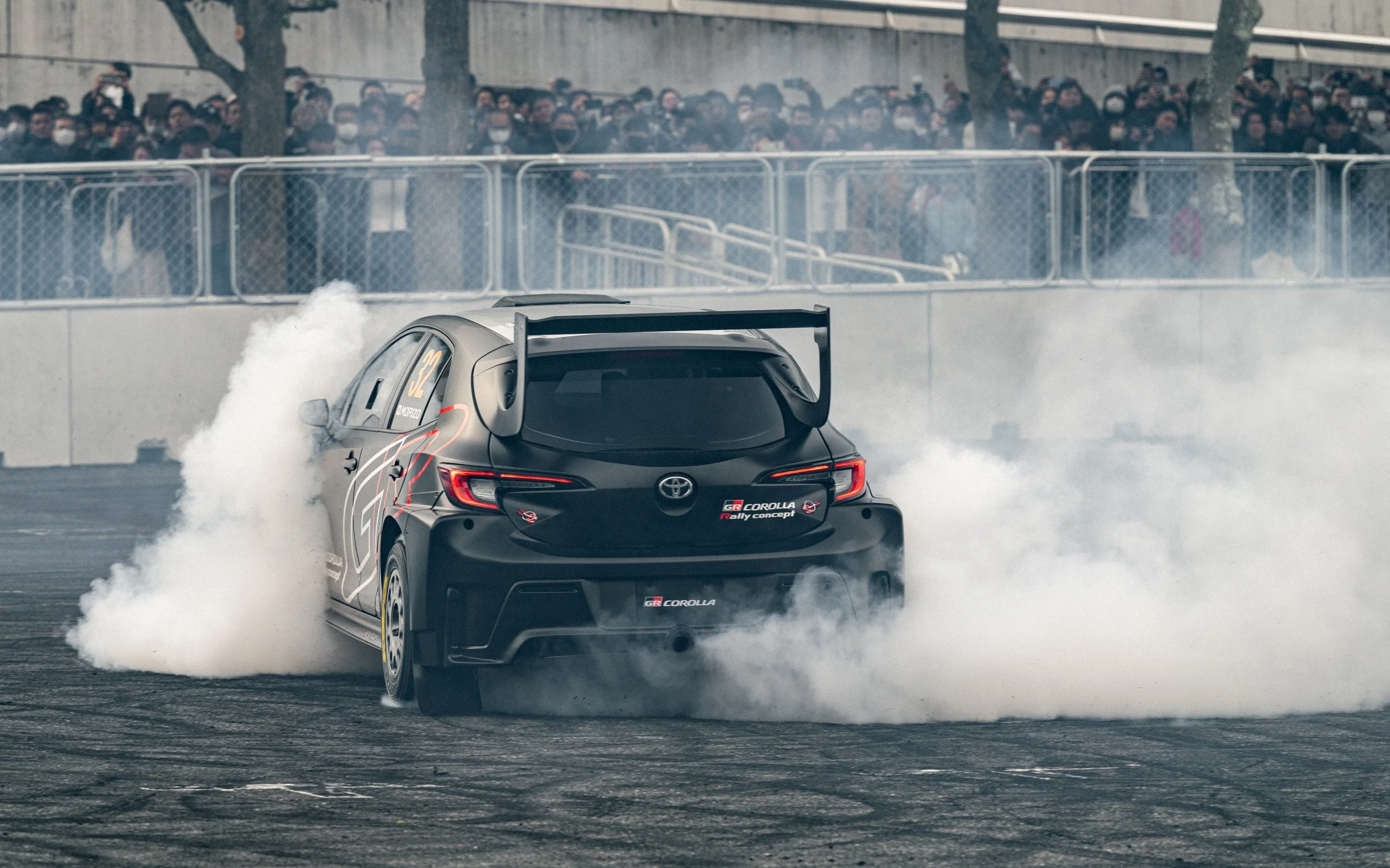 A black Toyota GR Corolla race car performing a burnout on a racetrack, with tire smoke and a crowd of spectators behind a protective fence.