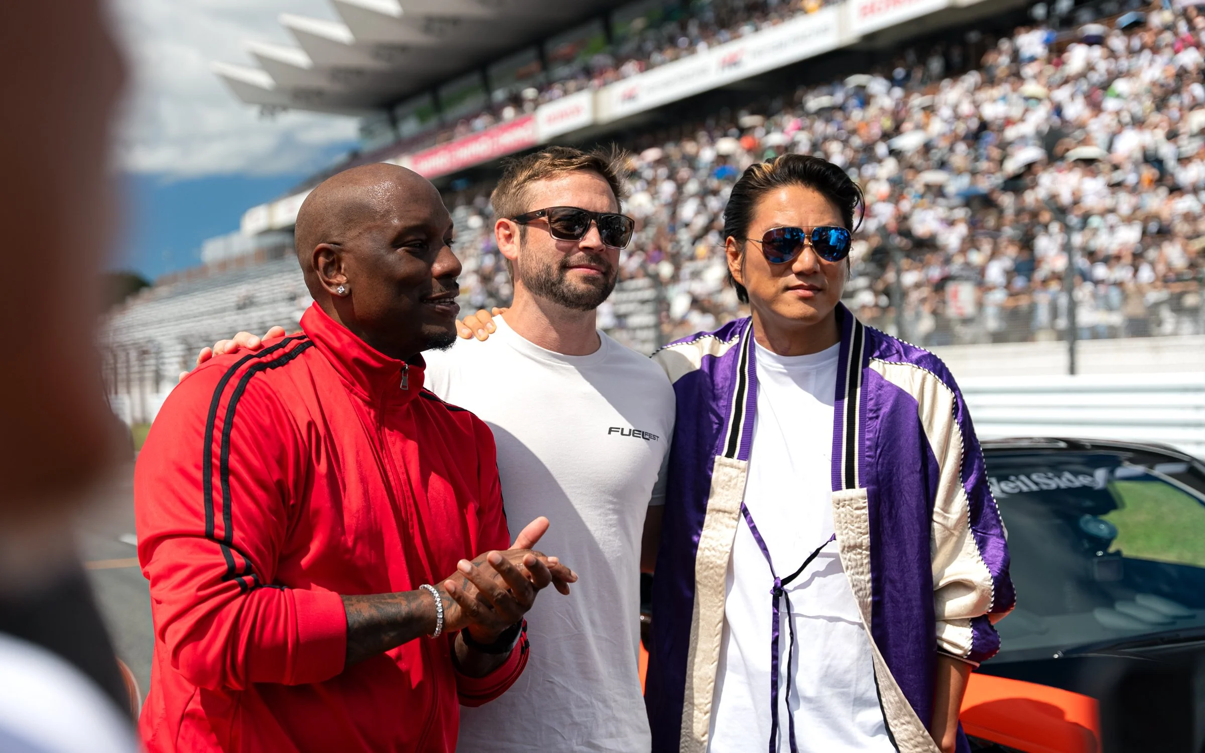 Three individuals standing on a race track with a crowded grandstand behind them during a sunny day, appearing to be at a motorsport event.