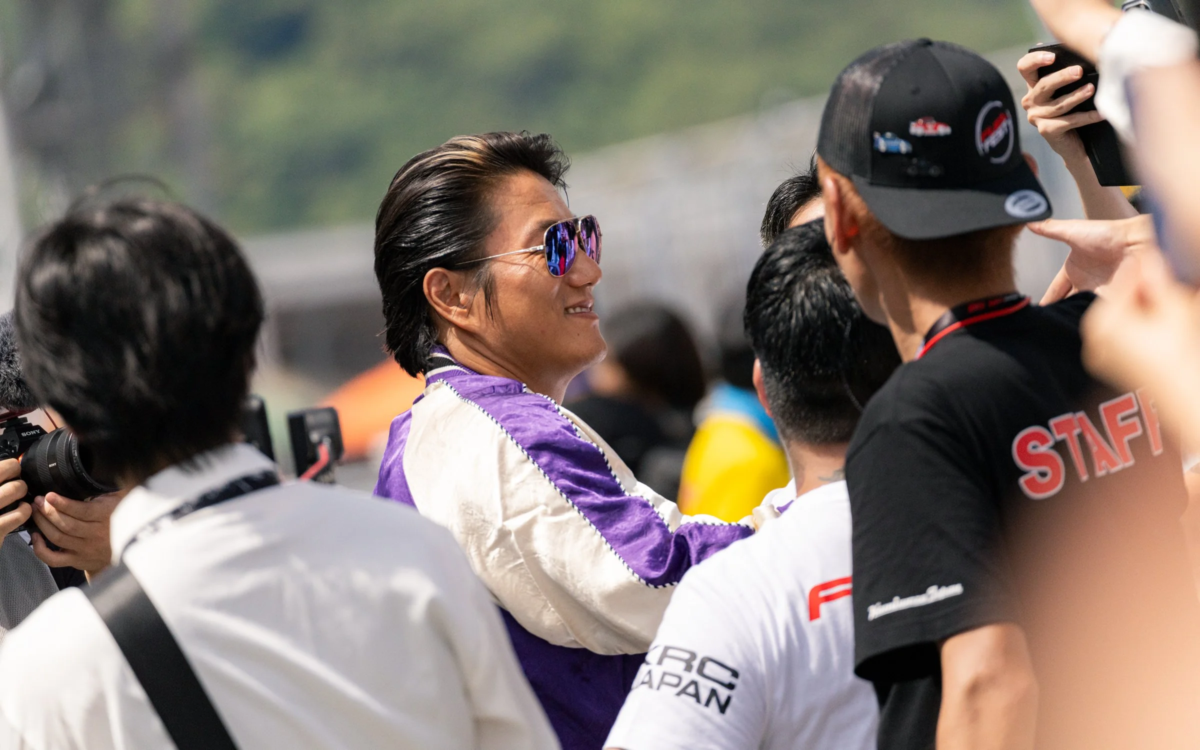 A person wearing sunglasses and a racing jacket interacts with team members at a racing event, with others taking photos and videos around him.