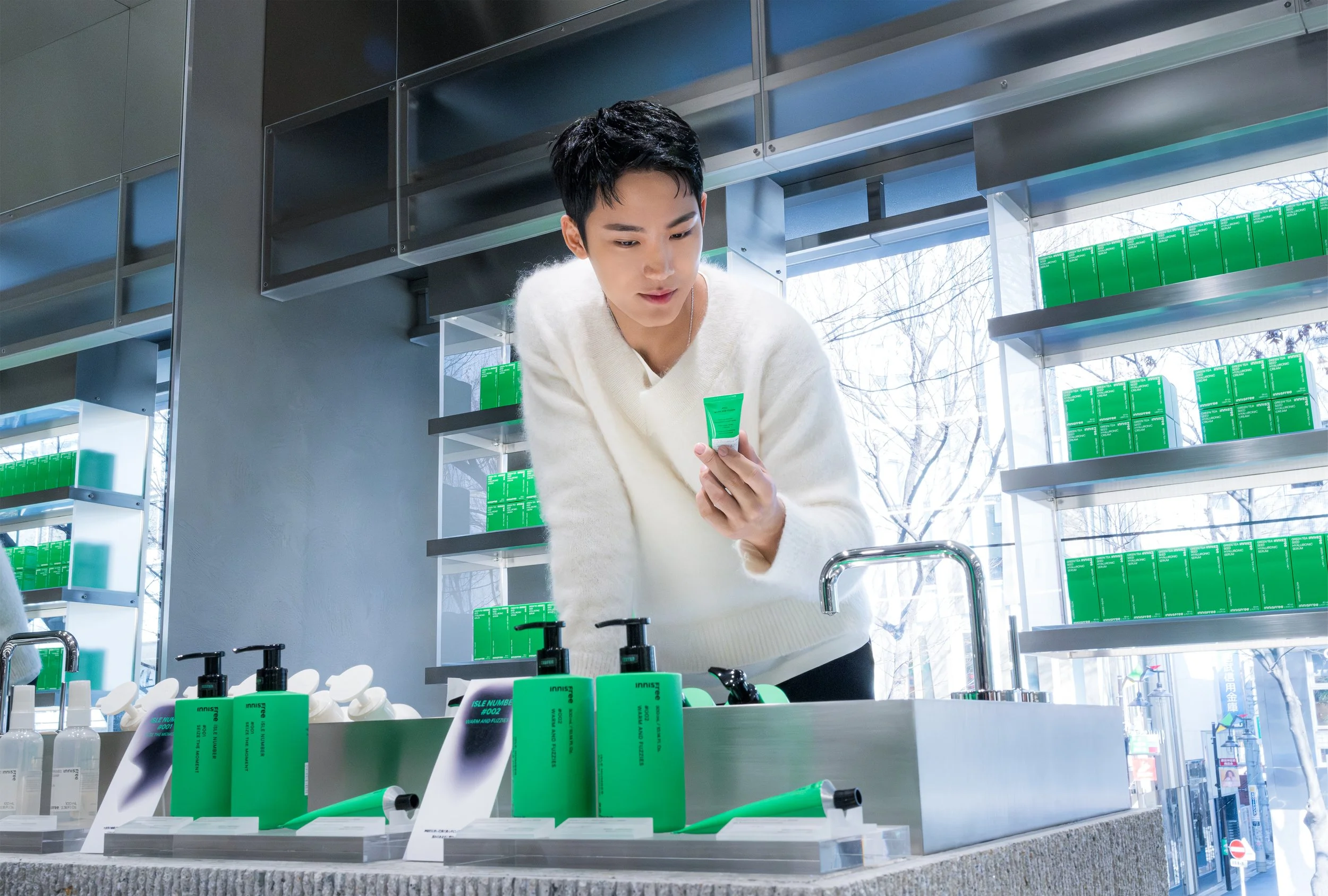 A young man in a white sweater shopping for skincare products at a store with shelves of green boxes and bottles, and a large store window in the background.