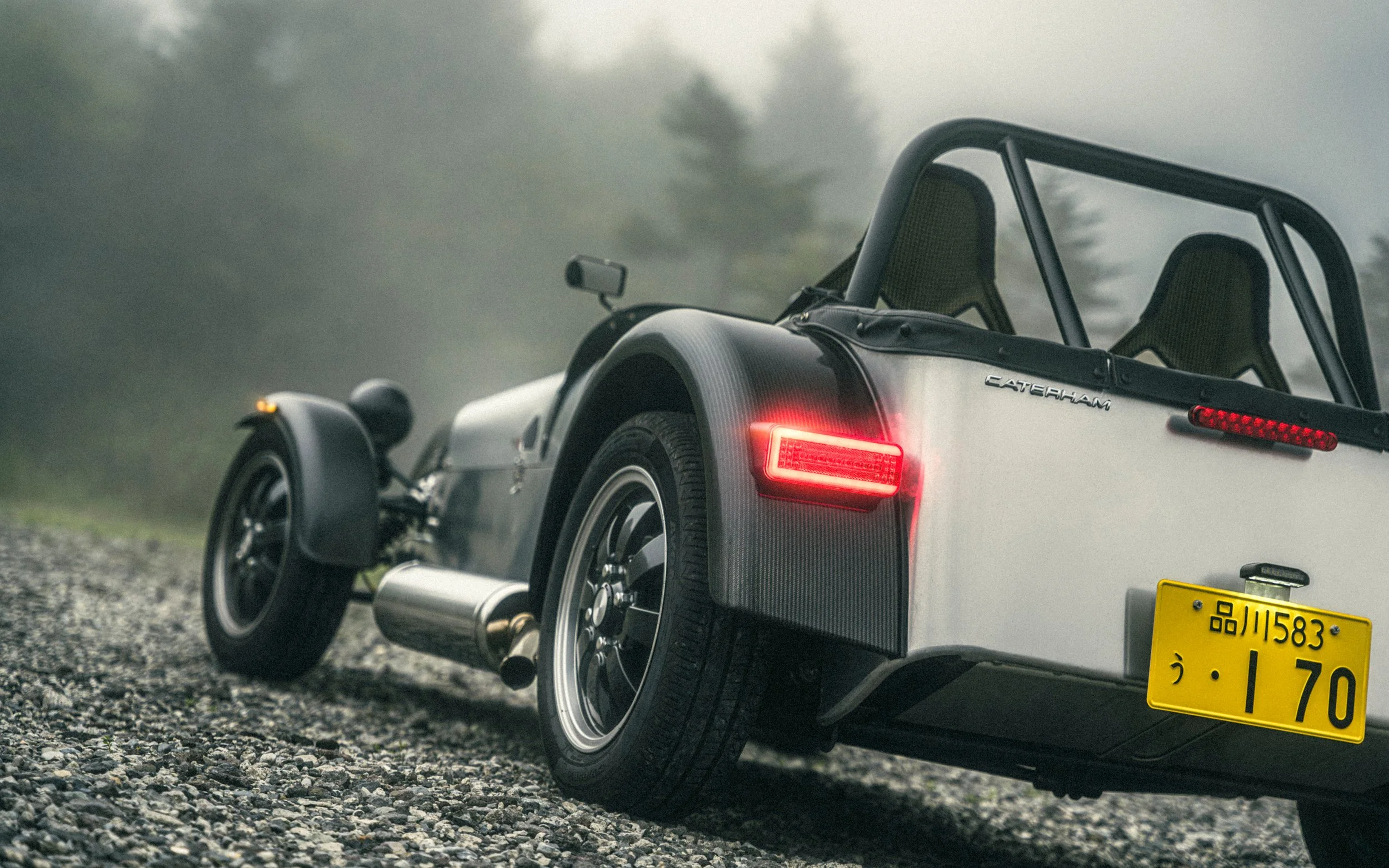 A black and white Caterham sports car with an open cockpit parked on a gravel surface in a foggy outdoor setting with trees in the background.