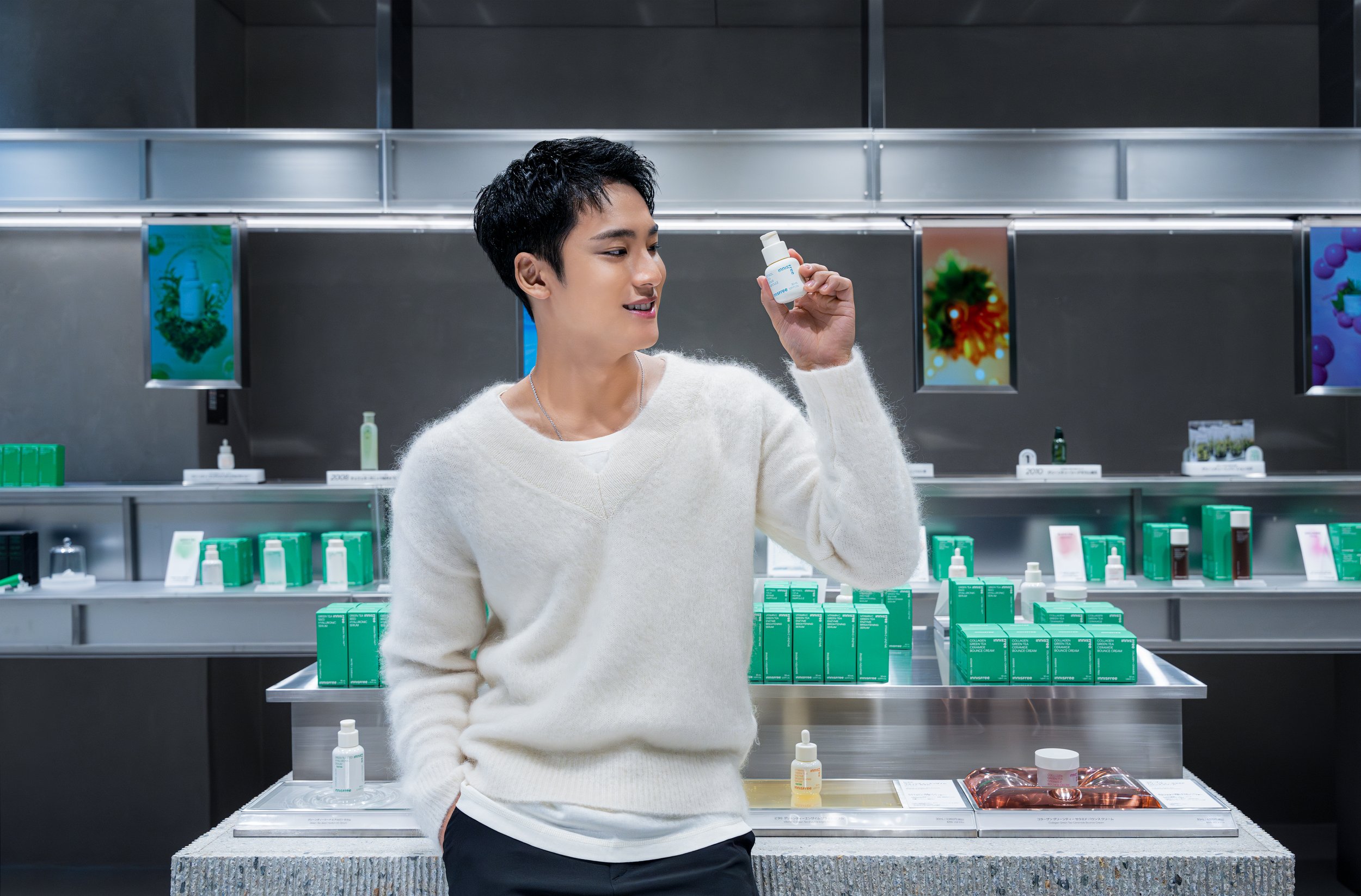 A young man with black hair and a white sweater shopping for skincare products in a store, holding a small white bottle and looking at it.