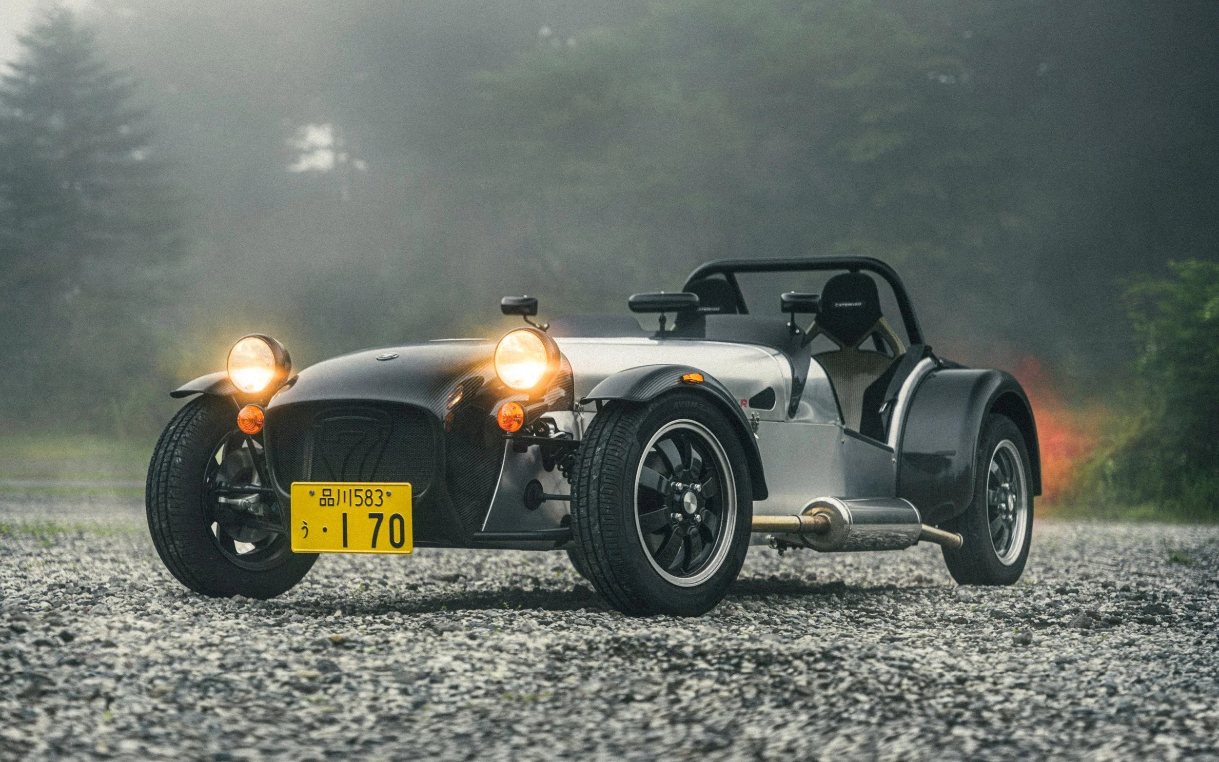 A vintage-style, open-wheel sports car with a black and silver body, round headlights on, and a Japanese yellow license plate, parked on a gravel surface with a misty, green background.