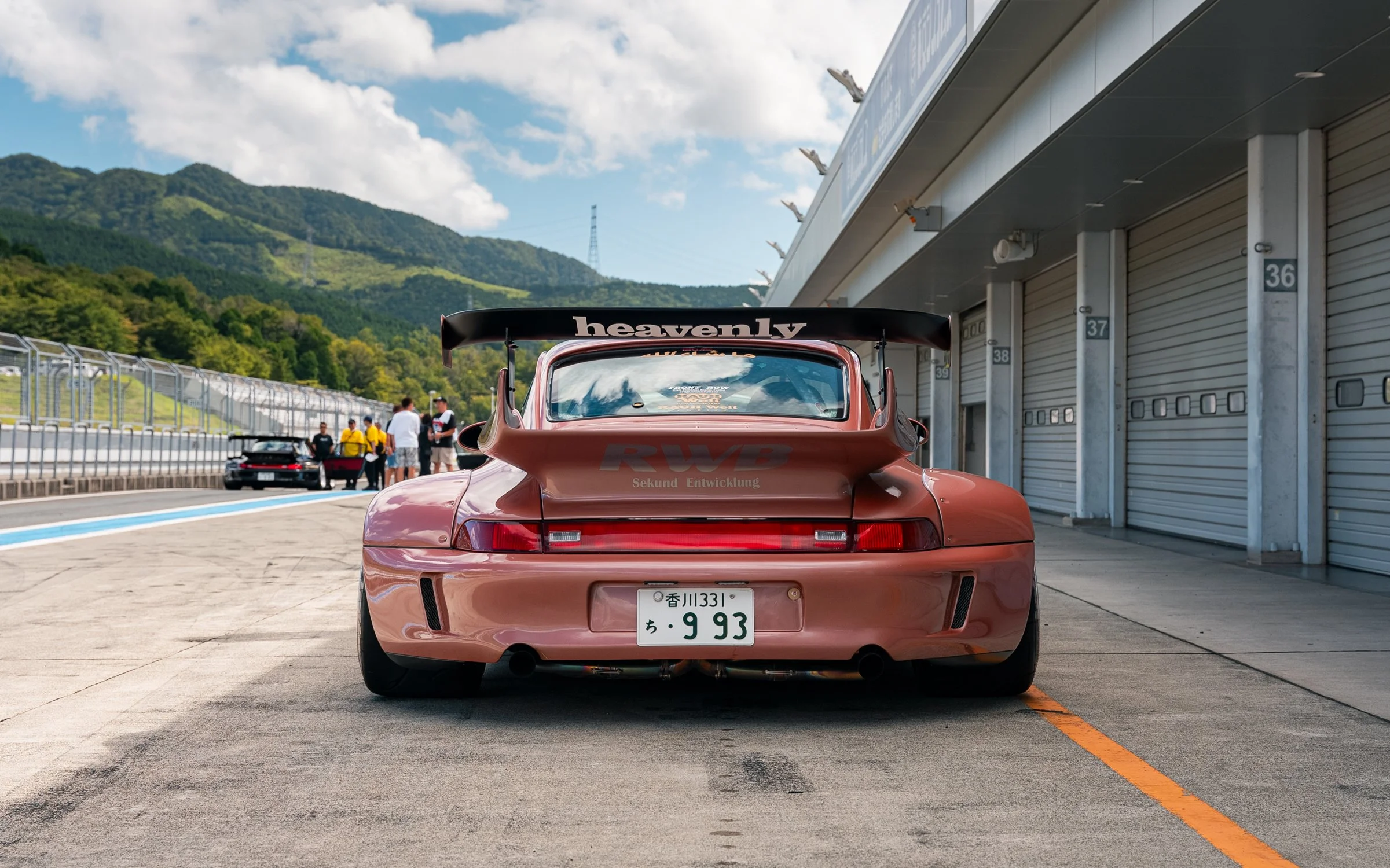 Rear view of a pink Porsche race car with a large rear wing labeled 'heavenly' parked in a pit lane at a race track, with mountains, a cloudy sky, and pit garages in the background.