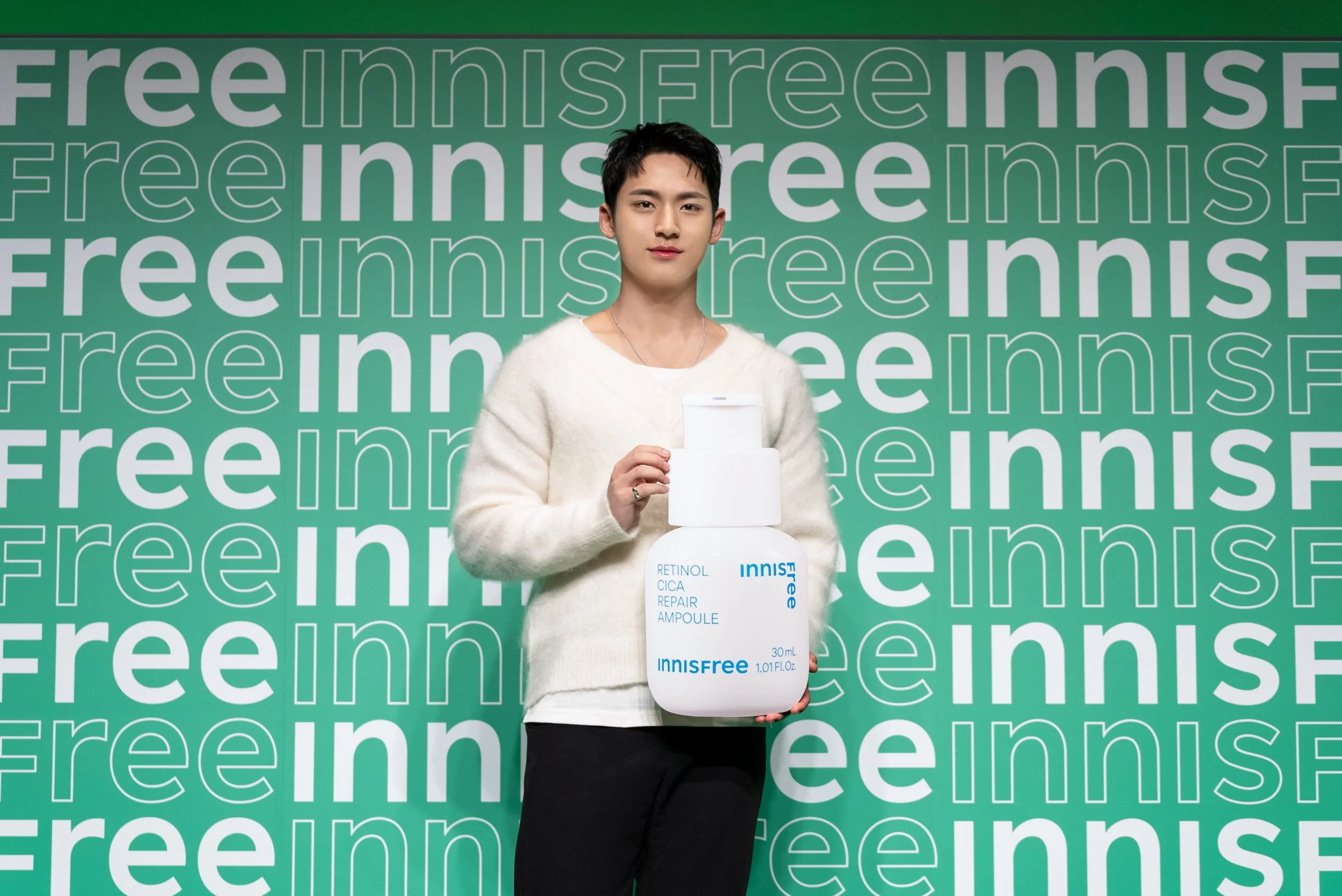 Young man standing in front of a green background with repeating white text, holding a large bottle of Innisfree Retinol Cica Repair Ampoule.