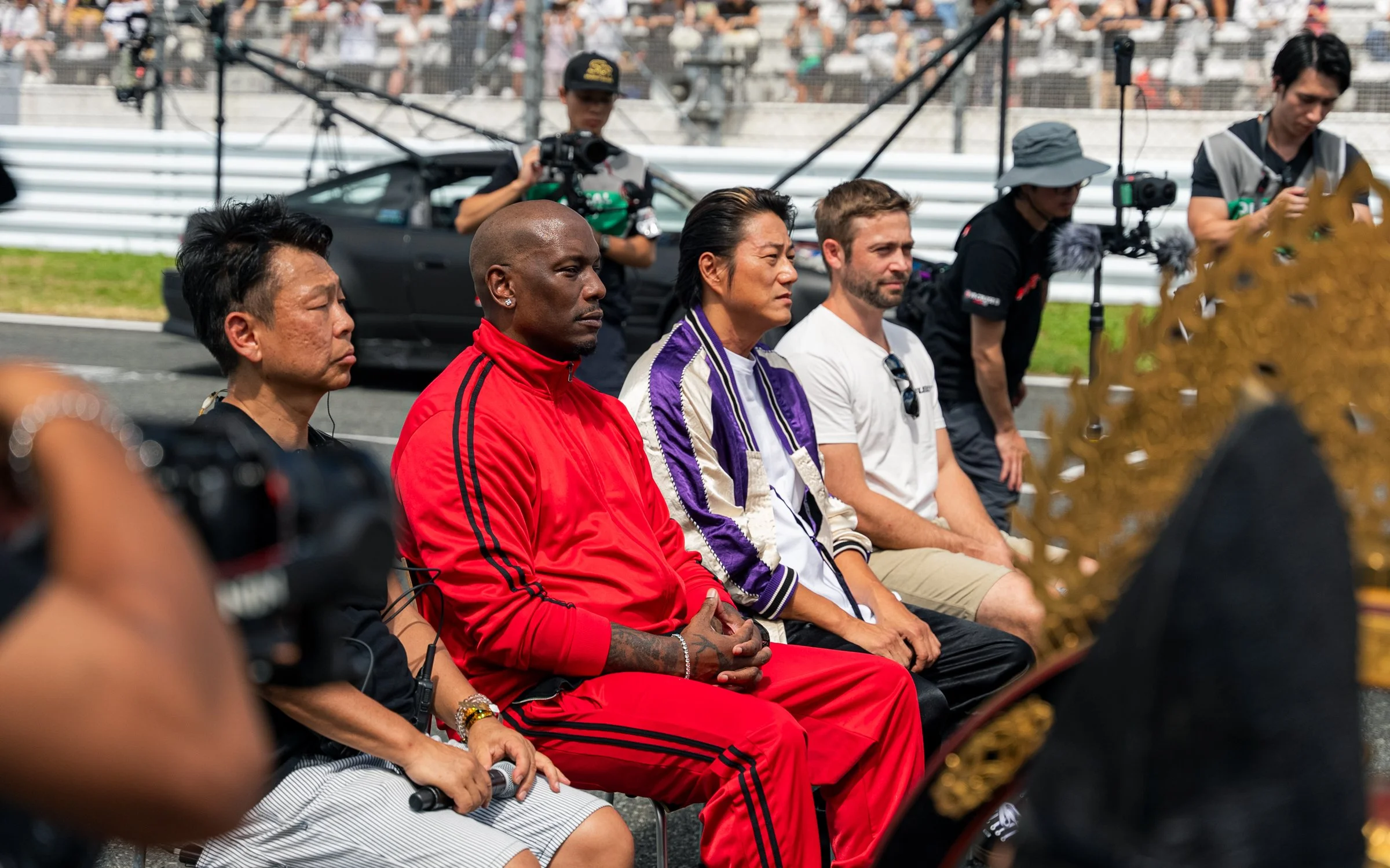Four people sitting on a row of chairs at a racing event, with journalists and cameras around them.