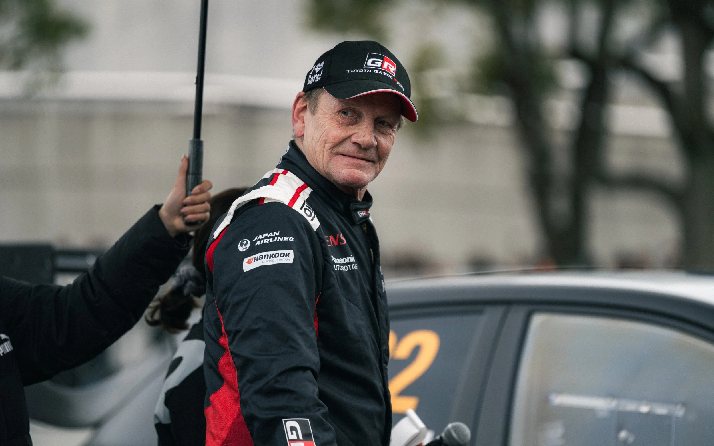 A race car driver in a black racing suit with red and white accents, wearing a black cap, standing next to a race car with the number 22, with a person holding an umbrella over him.