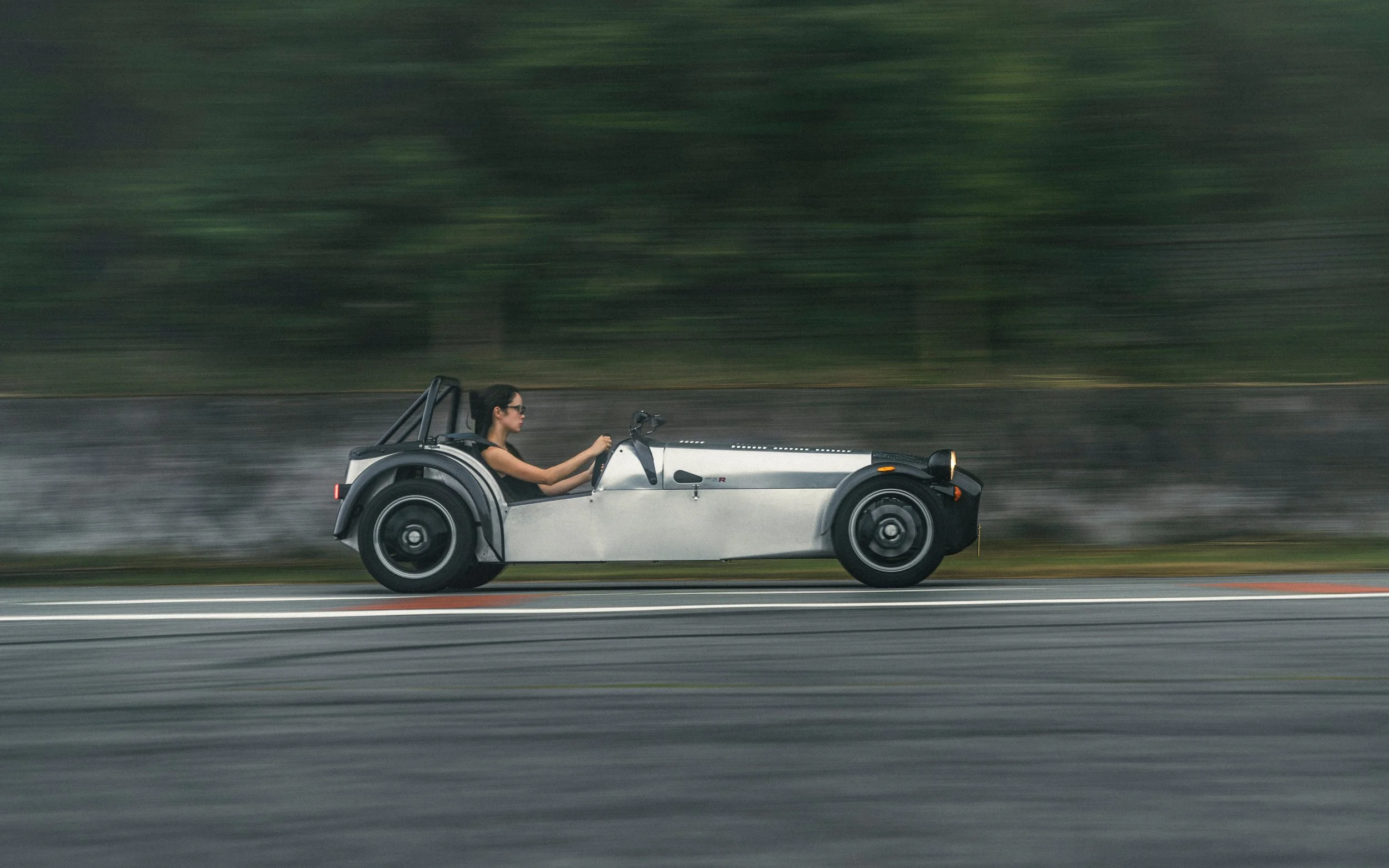 A woman driving a sleek, futuristic silver sports car on a racetrack with blurred trees in the background.