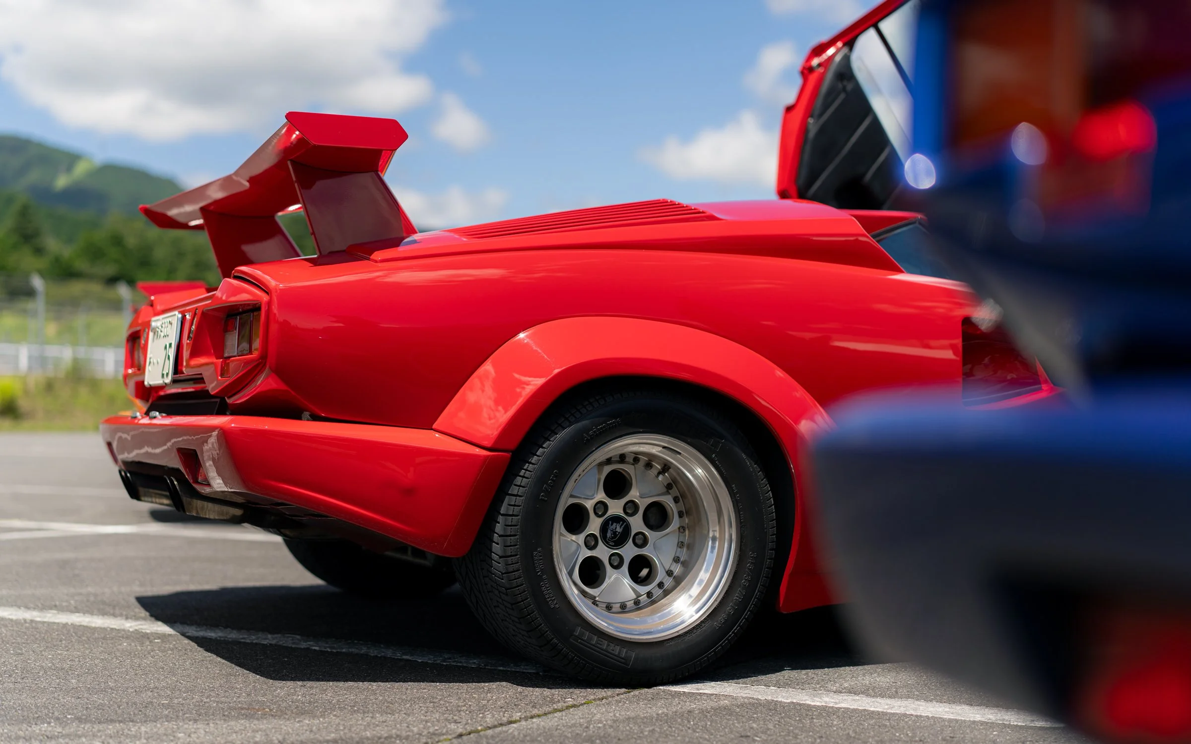 Side view of a red sports car with a prominent rear spoiler, parked on a race track or parking lot with mountains in the background and partly cloudy sky.