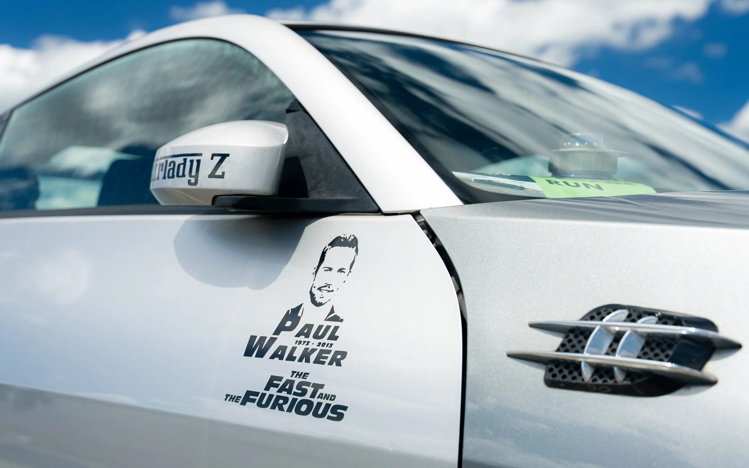 Close-up of a silver sports car with a decal of Paul Walker's face and the text "Paul Walker 1973 - 2013, The Fast and the Furious" on the door, a rearview mirror with a "Lady Z" sticker, and a vent on the side.
