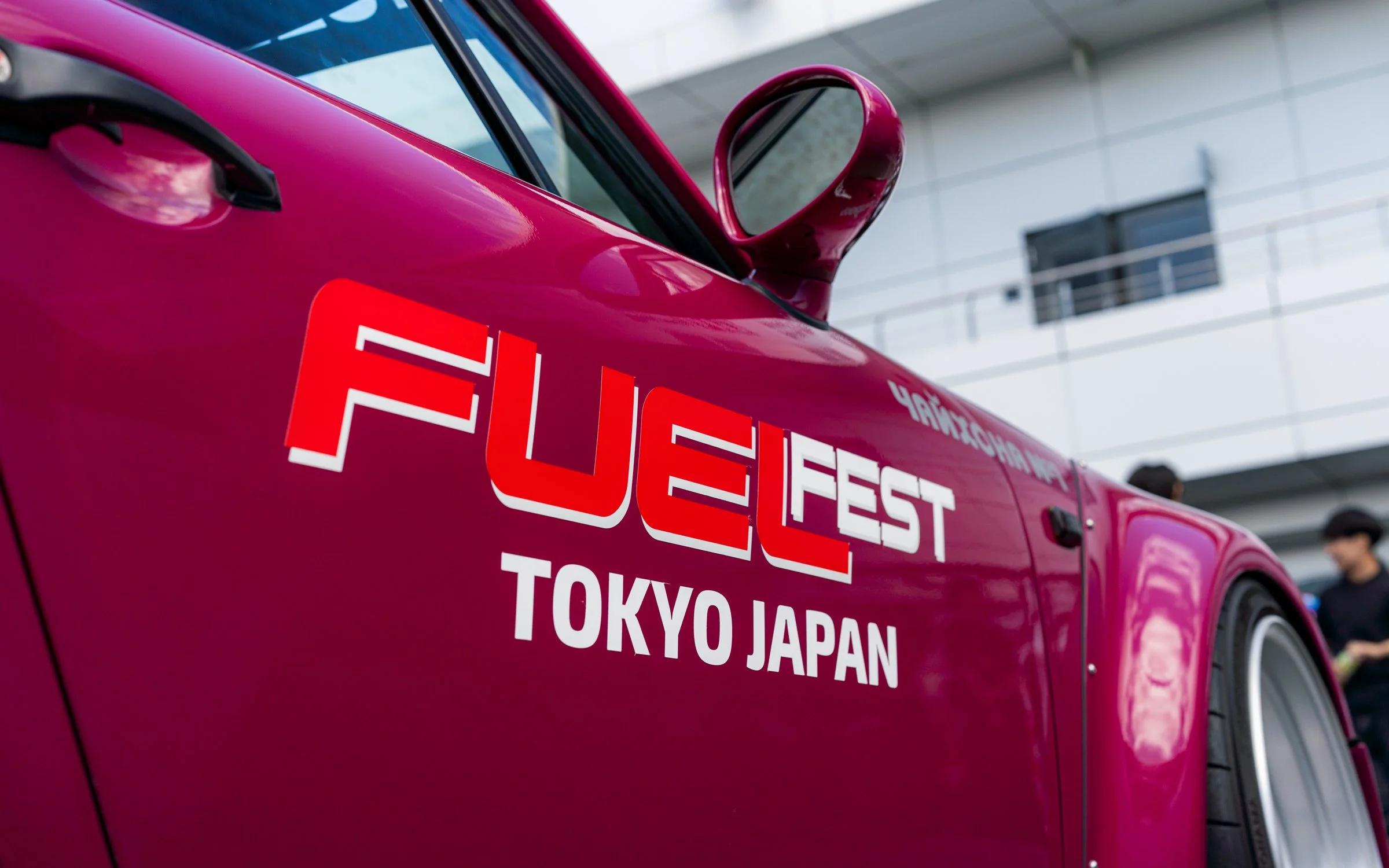 Close-up of a pink sports car with a 'FUEL FEST' and 'TOKYO JAPAN' decal, parked indoors with a building in the background and a person in black clothing nearby.