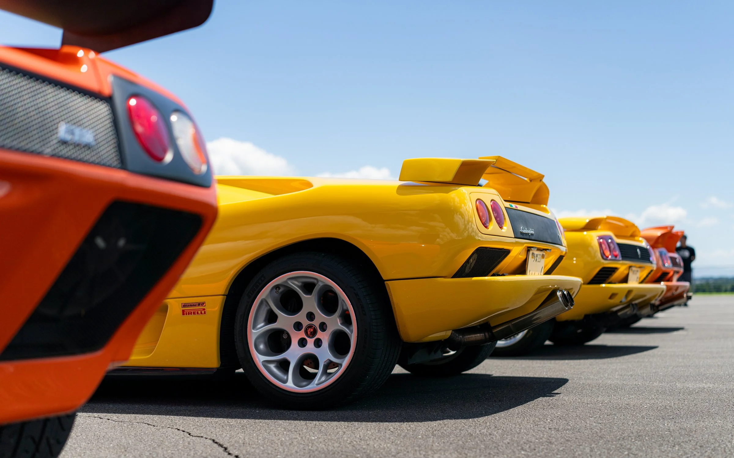 Line of yellow and orange sports cars parked on a tarmac under a partly cloudy sky.