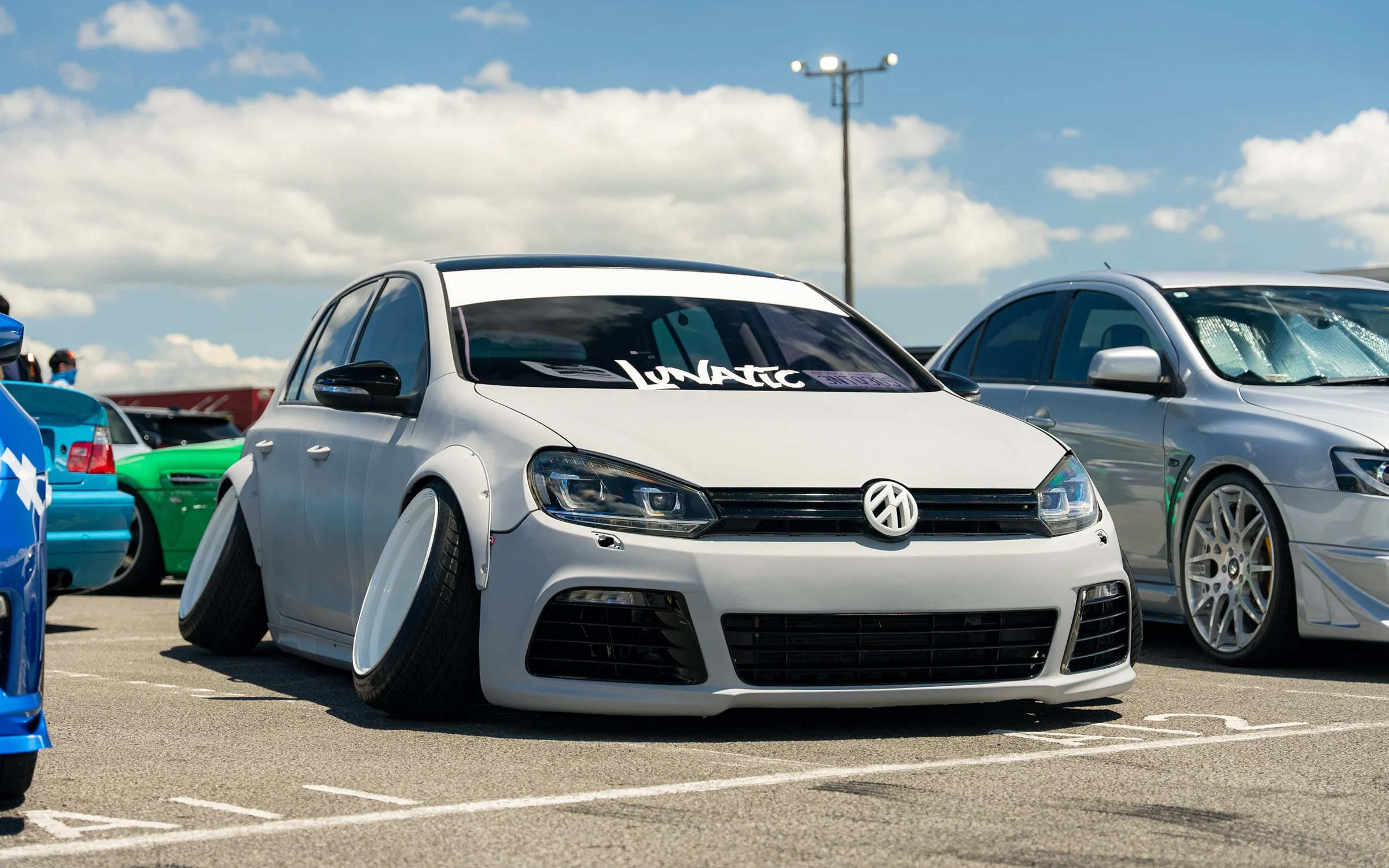 A modified white Volkswagen car with an aggressive stance, lowered suspension, and large wheels at a car show.