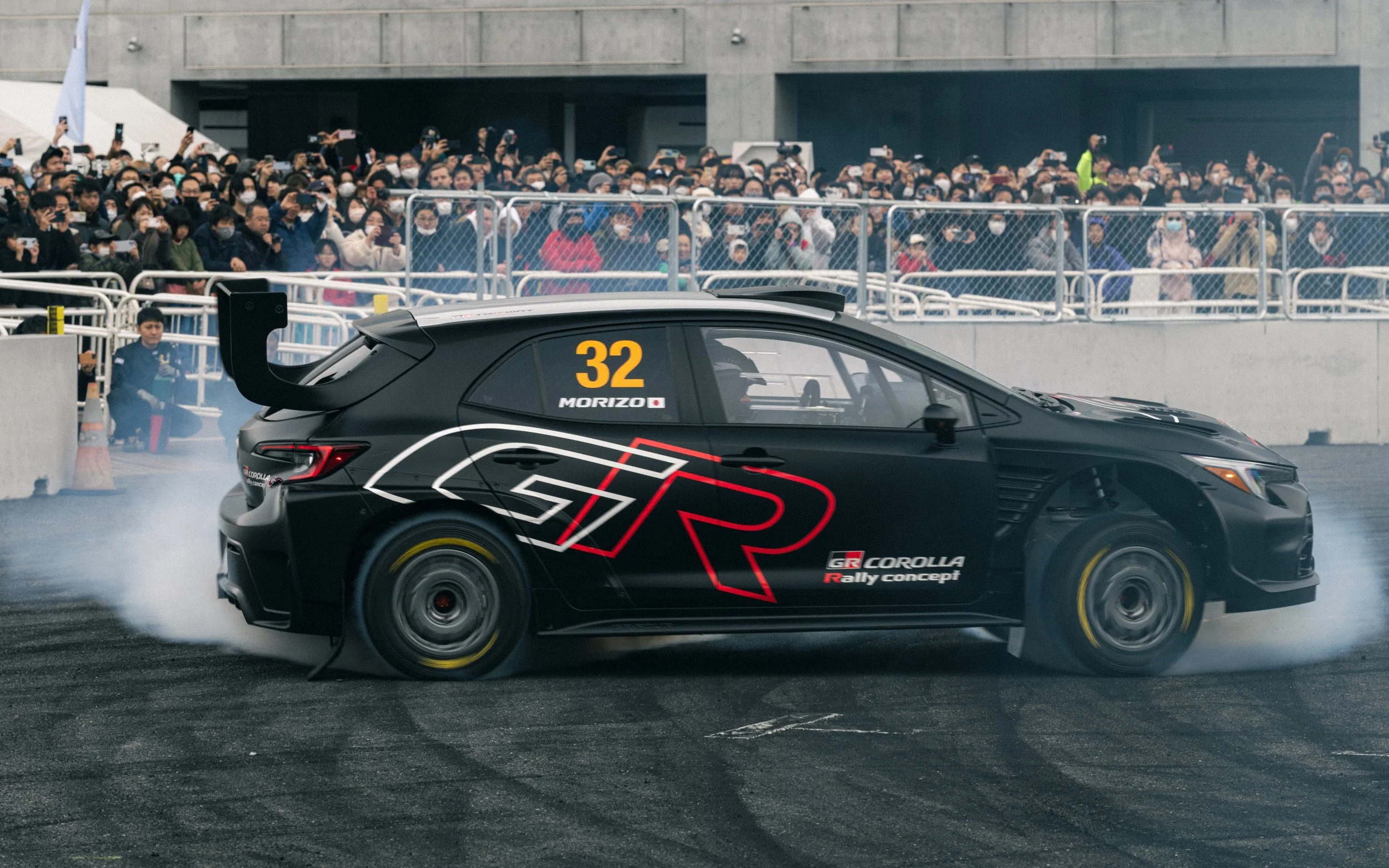 A black Toyota GR Corolla rally car with the number 32 and the name Morizo on the side, performing a burnout on a race track while a large crowd of spectators watches behind a fence.