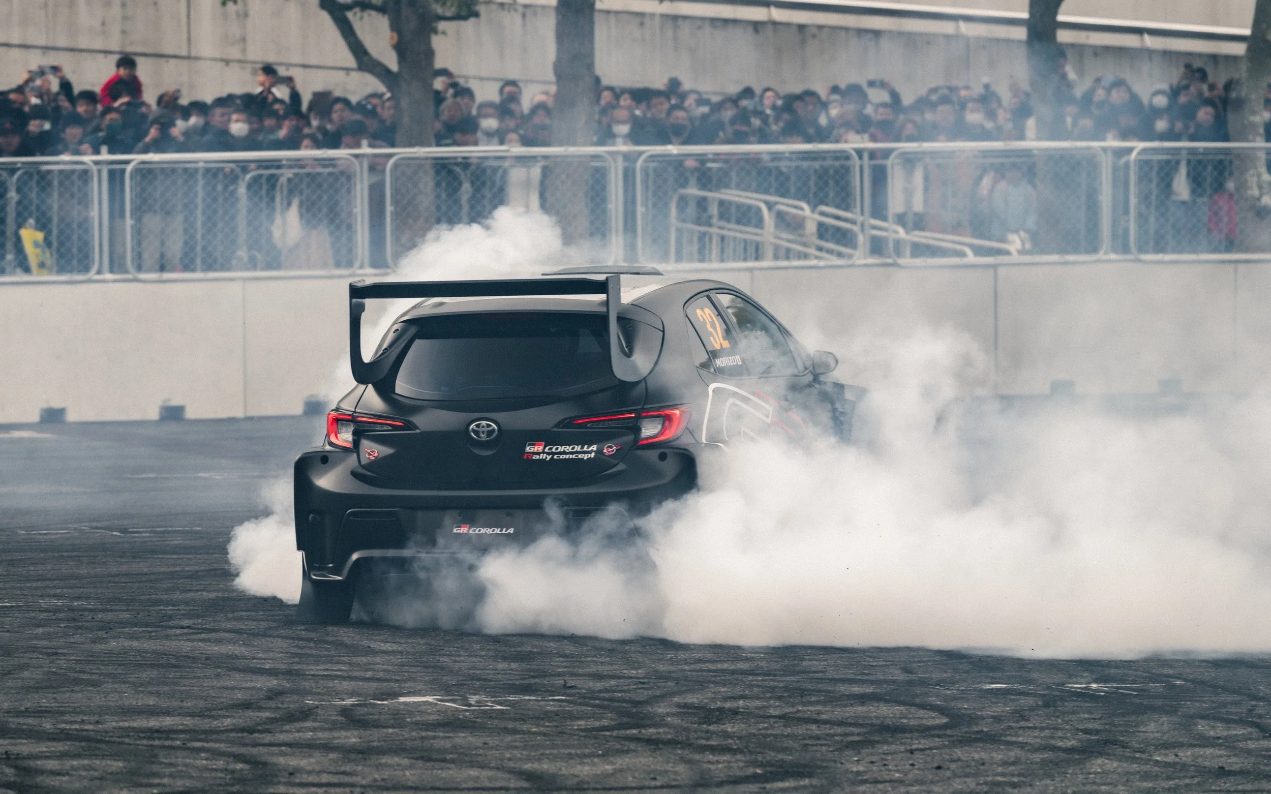A black race car executing a burnout on a track with smoke billowing from the tires, a crowd of spectators watching from behind a fence, and trees in the background.