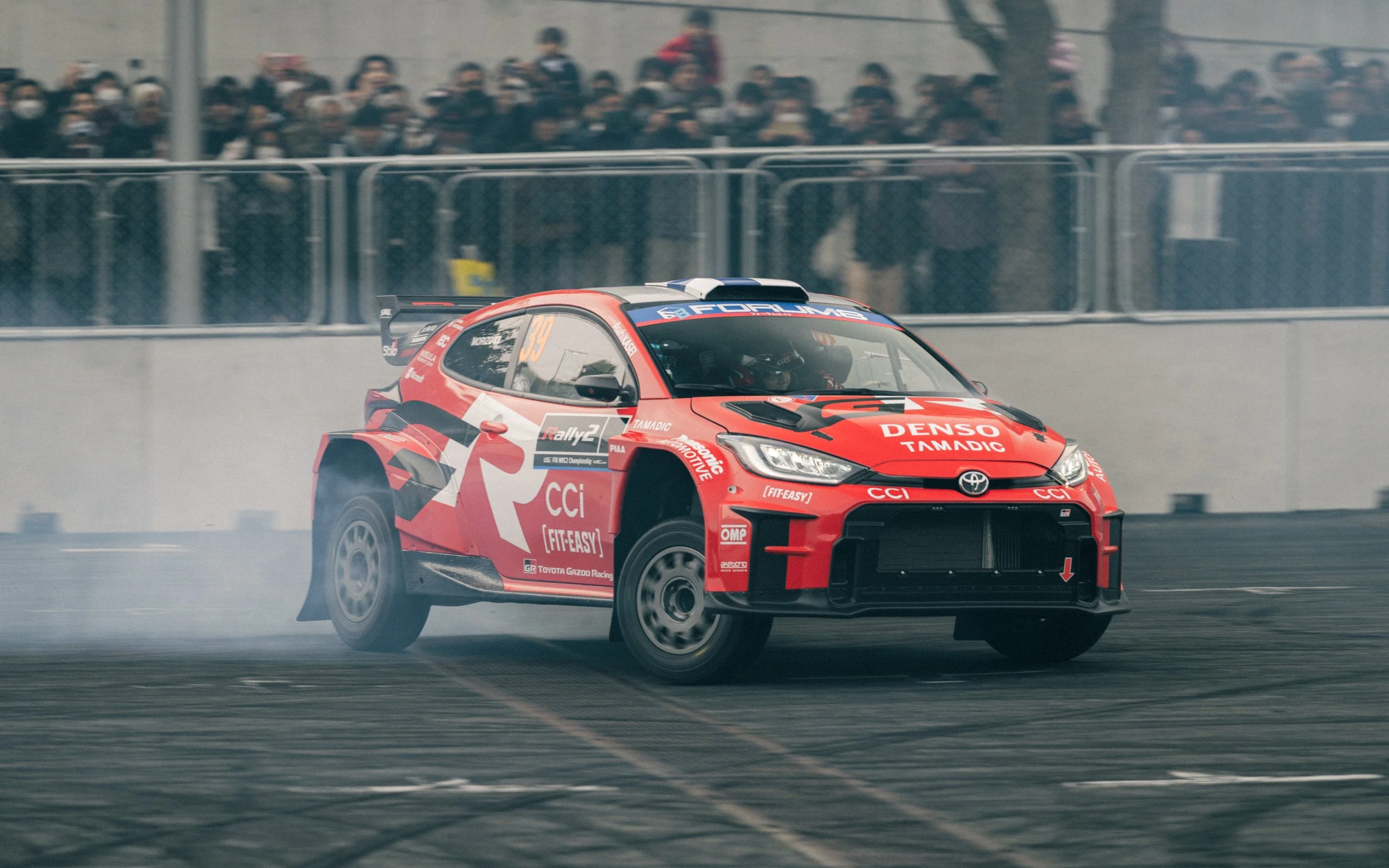 A red Toyota rally car drifting on a race track with smoke coming from the tires, and a crowd of spectators behind a fence in the background.