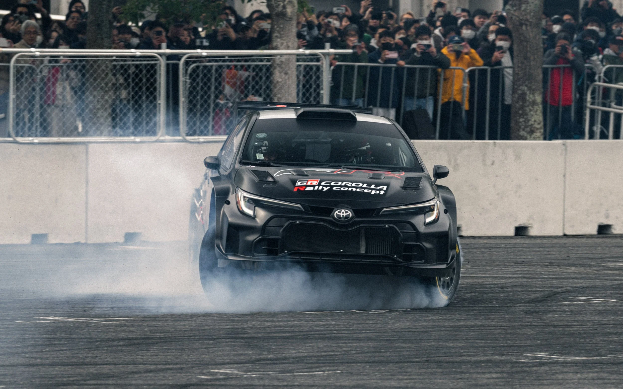 A black Toyota GR Corolla rally car drifting on a racetrack, with smoke coming from the rear tires and a crowd of spectators behind a fence watching.