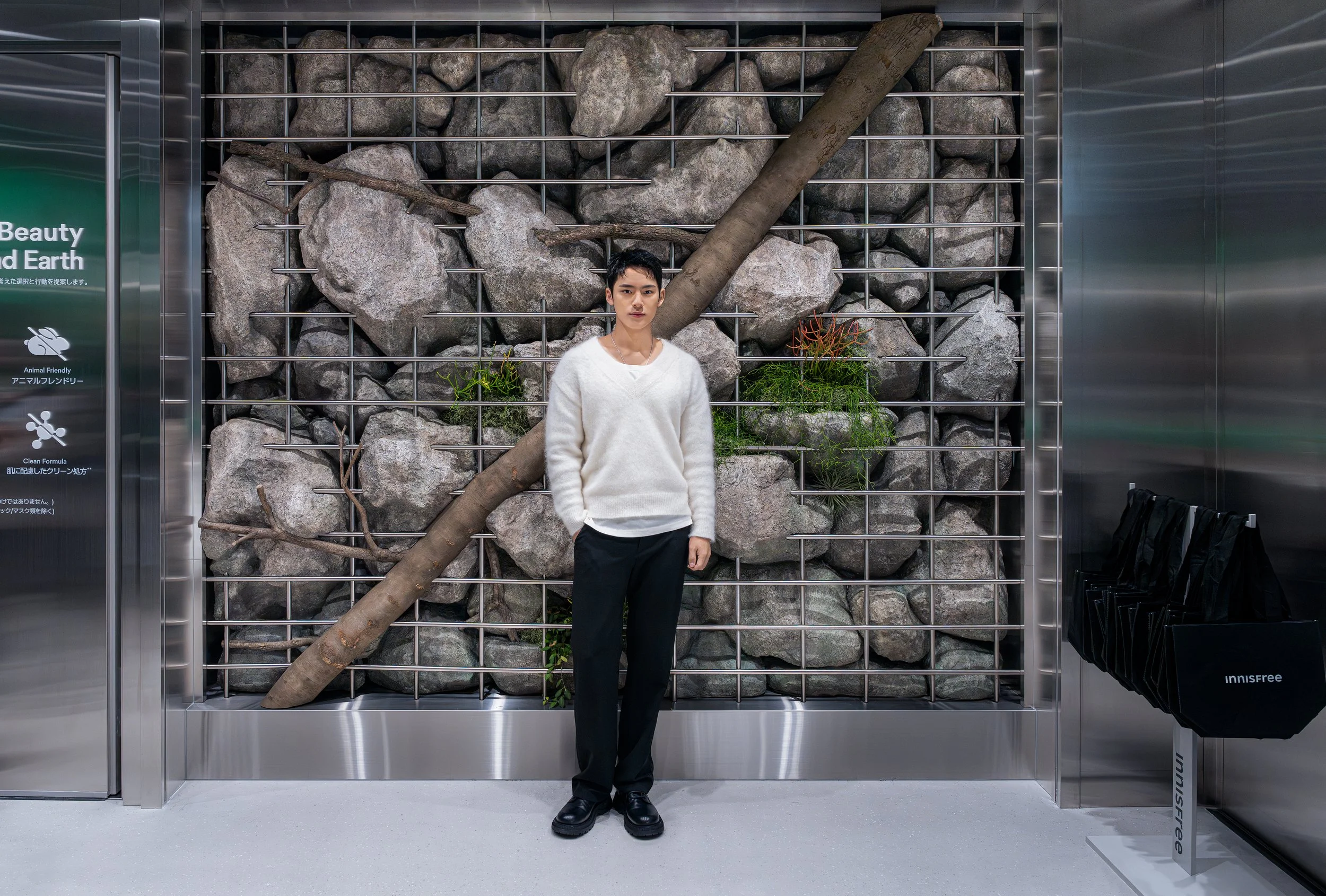A young man standing in front of a decorative wall with large rocks, metal grid, and plants, inside a modern building.