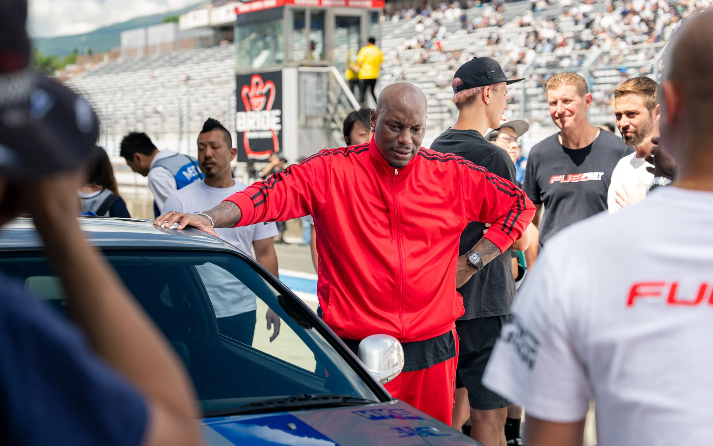 A man in a red sports jacket and black cap standing near a race car, surrounded by a group of people at a race track.