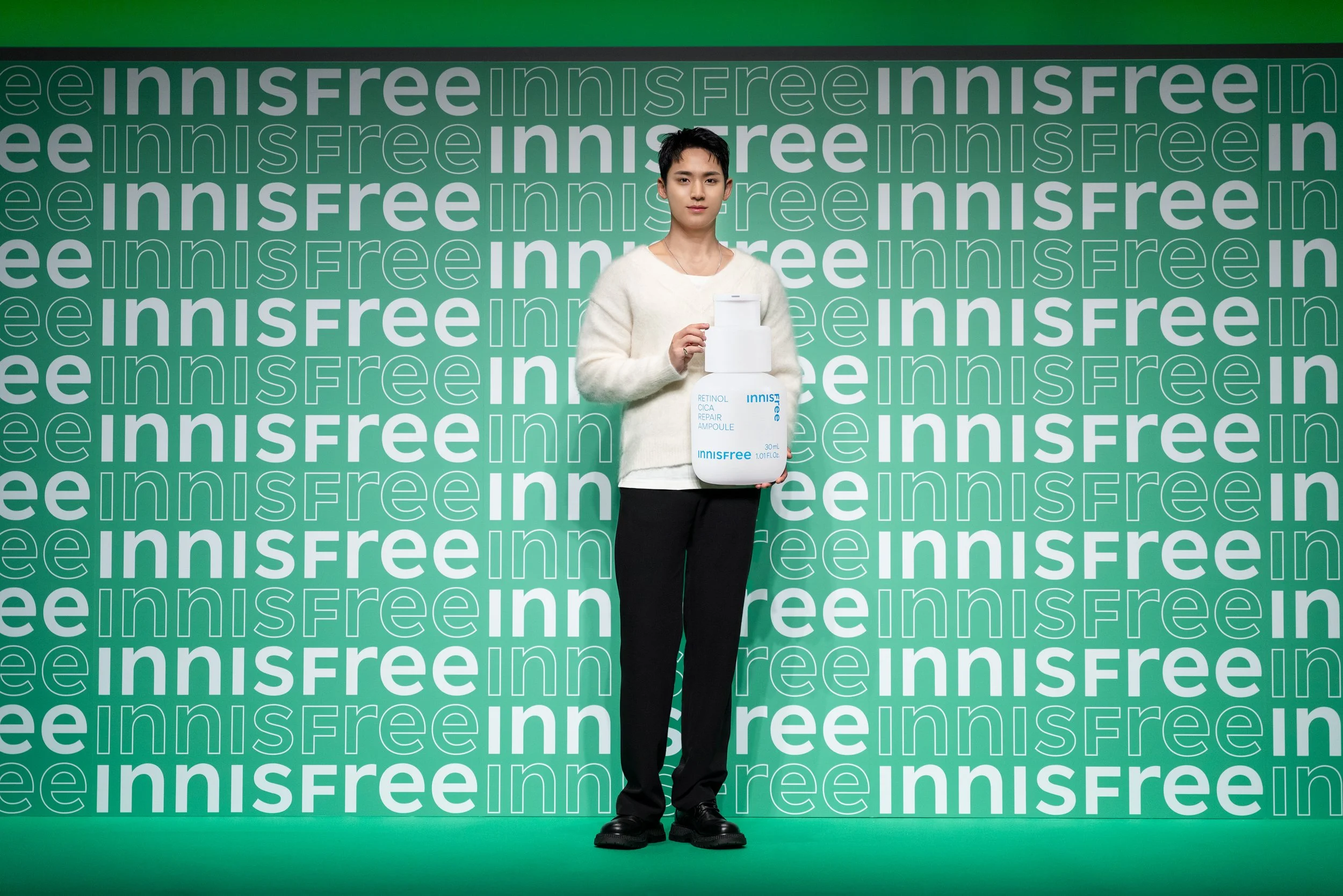 A woman standing in front of a green and white background with the words 'innisfree' repeated. She is holding a large, white bottle of Innisfree skincare product labeled 'Retinol Cica Repair Ampoule'.